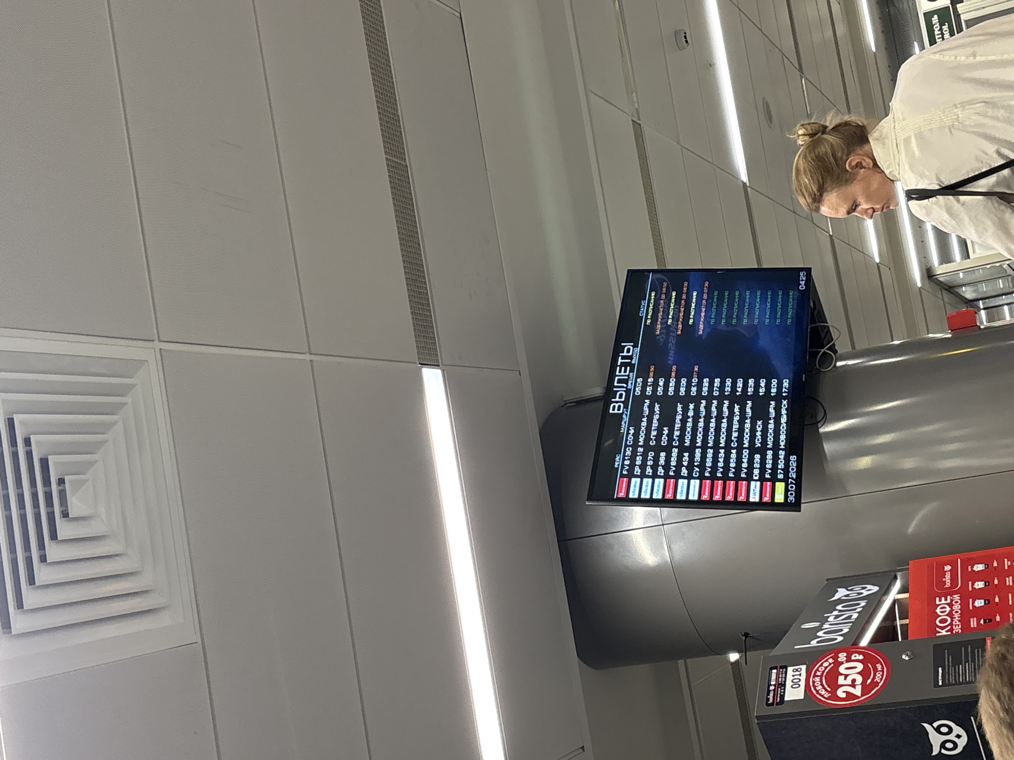 An indoor, eye-level, medium shot captures a departure information board at an airport. The board displays flight information with columns for time, flight number, destination, and status. A woman with blonde hair pulled back in a bun is visible from the back, to the right of the board, facing away from the camera. She is wearing a white collared shirt and a black strap is visible over her shoulder. A coffee stand, branded "barista," is situated to the left of the departure board, featuring red signage and an owl logo. The ceiling of the airport, with its ventilation system and fluorescent lighting, is prominent in the upper portion of the image. The departure board shows flight information, including a date stamp of "30.07.2026" and a time of "04:25". The context suggests this scene is within the Bolshoye Savino airport in Russia.