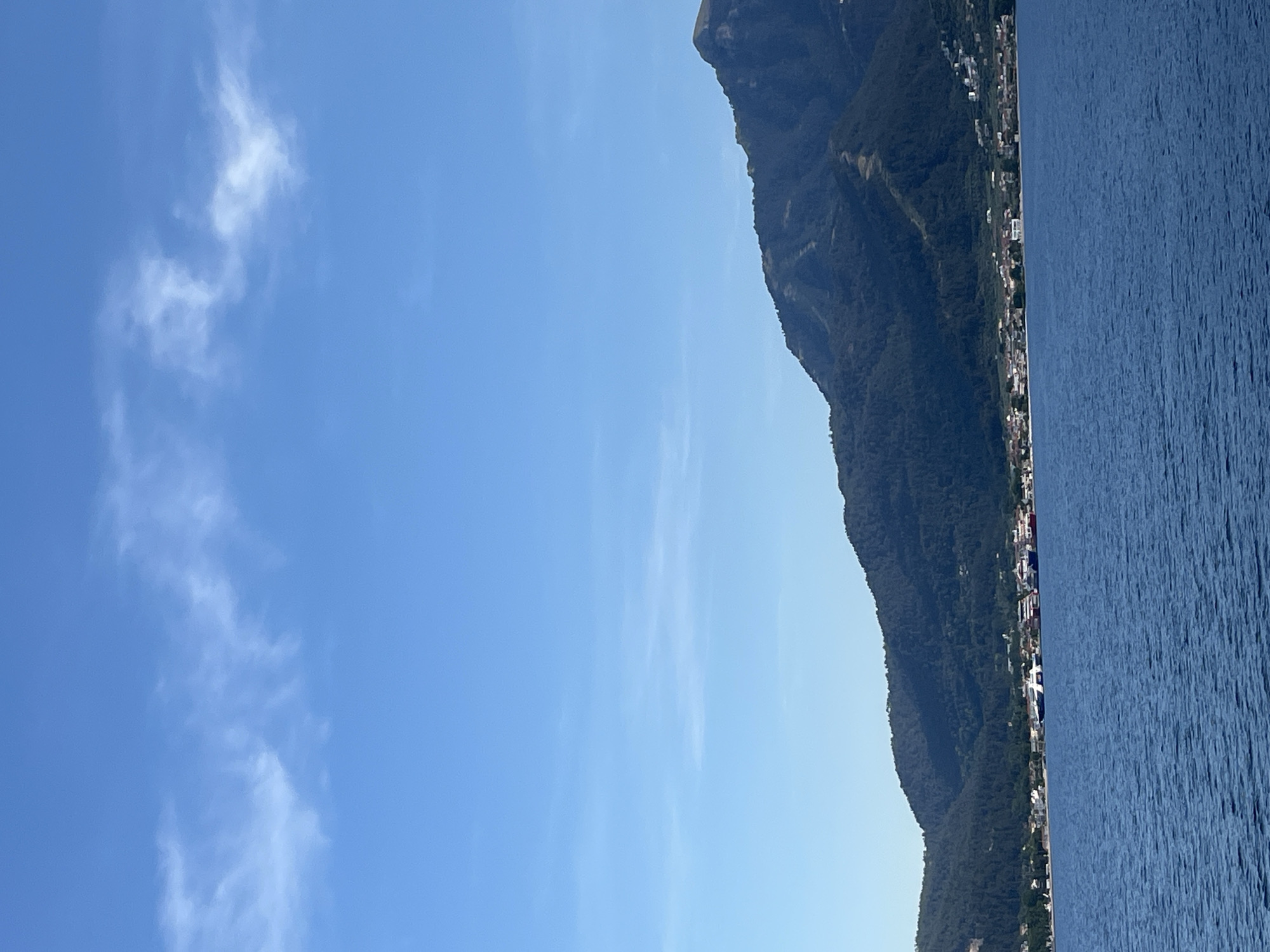 A vertical, eye-level, medium-shot captures a coastal scene with a clear blue sky and a body of water in the foreground. The sky is a bright cerulean, with sparse, wispy white clouds scattered across the upper half. A chain of dark green, tree-covered mountains dominates the background, sloping down towards the coastline. Nestled along the base of the mountains is a town with white buildings and red-tiled roofs, interspersed with trees. Several ferries, including a large white and blue vessel and two red-hulled ferries, are docked at the town's harbor. The water in the foreground is a deep, rippling blue, reflecting the light of the sky.