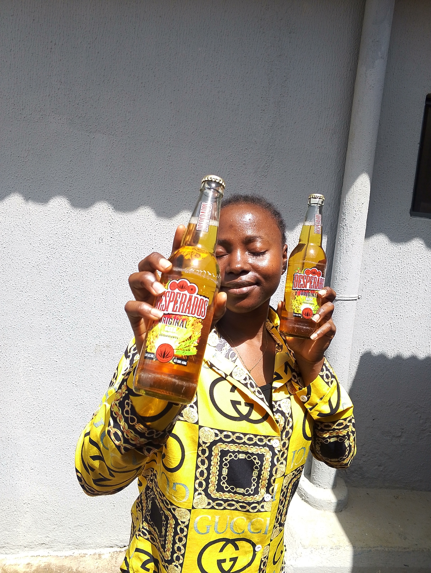 A young woman with dark skin and closed eyes is holding two bottles of Desperados Original beer in front of a gray, textured wall. She is wearing a bright yellow shirt with a bold black and gold pattern, featuring interlocking G symbols and chain designs. Sunlight casts distinct shadows on the wall, creating a wavy pattern. The woman is positioned in the center of the frame, with the bottles held at chest level. One bottle is in her right hand, closer to the viewer, and the other is in her left hand, slightly further away. The background appears to be an outdoor setting, possibly in a residential or public area in Jos, Nigeria. The overall lighting suggests a bright, sunny day.