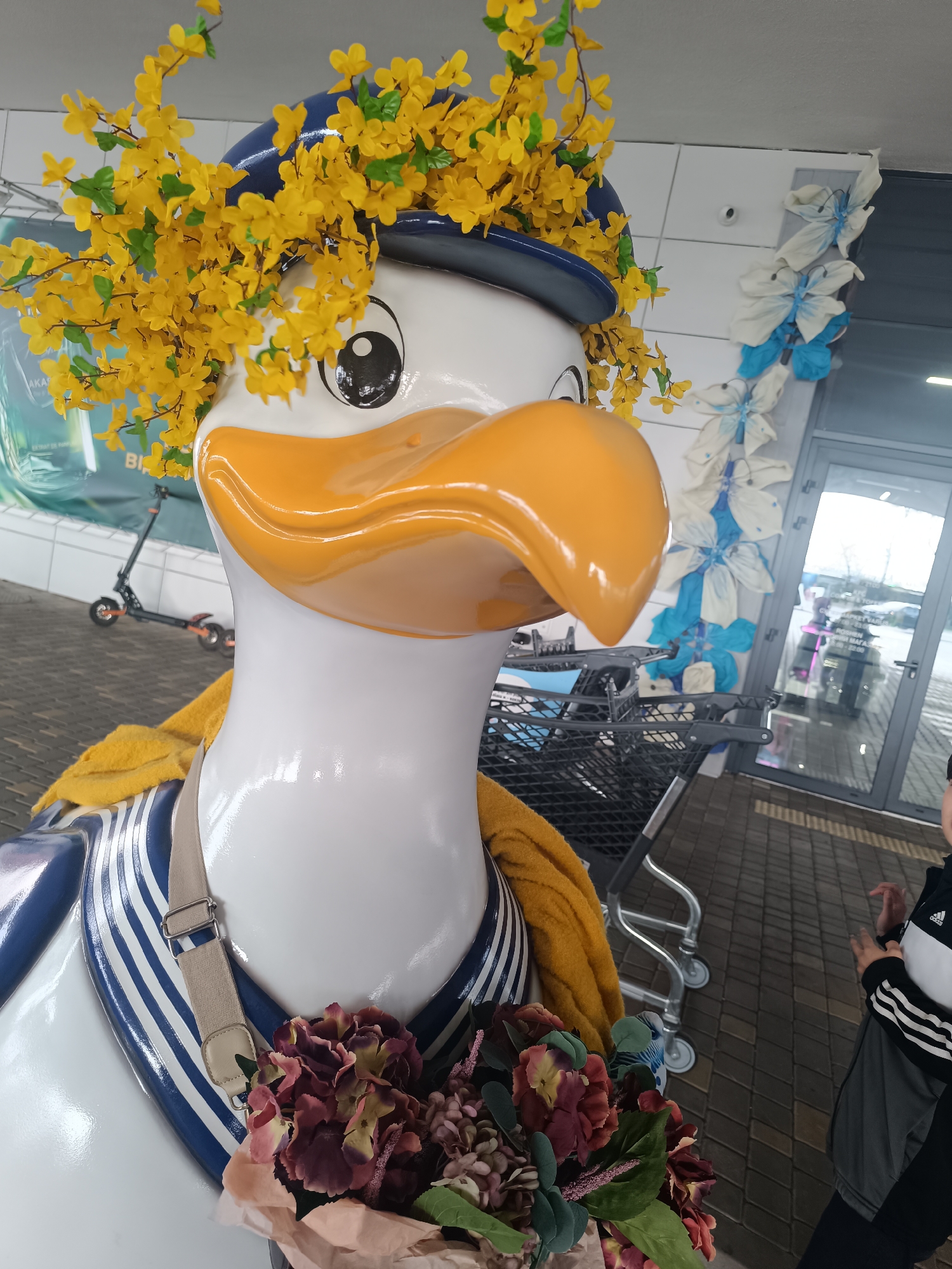 A large, white statue resembling a cartoon duck, possibly intended as a mascot or decoration, stands in what appears to be a commercial entrance area. The duck wears a blue and white striped sailor-style collar and a dark blue hat adorned with a profusion of bright yellow artificial flowers. A yellow fabric shawl is draped around its neck, and a beige strap with a buckle crosses its chest. The statue holds a bouquet of artificial flowers in shades of deep red, purple, and pink.
The setting features a tiled ground and a glass entrance door in the background. To the right of the statue, part of a person wearing a black, grey, and white Adidas jacket is visible. A shopping cart is partially seen behind the duck. On a wall next to the glass door, decorative light blue and white butterfly shapes are arranged vertically. Further back on the left, a black and orange electric scooter is parked near a wall displaying a large green and blue advertisement banner.
The scene is lit by ambient daylight, suggesting it is daytime. Text on the glass door, though somewhat blurry, appears to indicate store hours or information, possibly reading "РОБОТА МАГАЗИНУ" (store hours) and "10:00 - 22:00", confirming the location as a shop entrance. The overall impression is a cheerful and decorated space, possibly a storefront, using the duck statue as an inviting display.