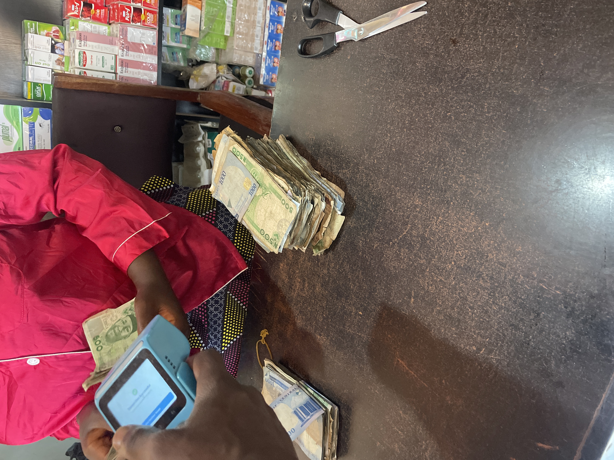 The image depicts a scene on what appears to be a counter in a shop. The counter is dark brown with a textured surface. On the counter are stacks of Nigerian Naira banknotes of various denominations, including 500 and 1000 Naira notes. A pair of scissors is lying on the counter towards the upper right of the frame.
A person, possibly a shopkeeper, wearing a red shirt and a patterned fabric on their lap is holding a mobile payment terminal. They are also holding some Naira banknotes. The terminal screen shows a green checkmark and text, indicating a successful transaction.
In the background, shelves are visible filled with various pharmaceutical products, including boxes labeled "FLU-J", "Exiplon", "Novalyn", "COFLIN", and "plumal".
There is no explicit information on the city of Durimi, Nigeria.