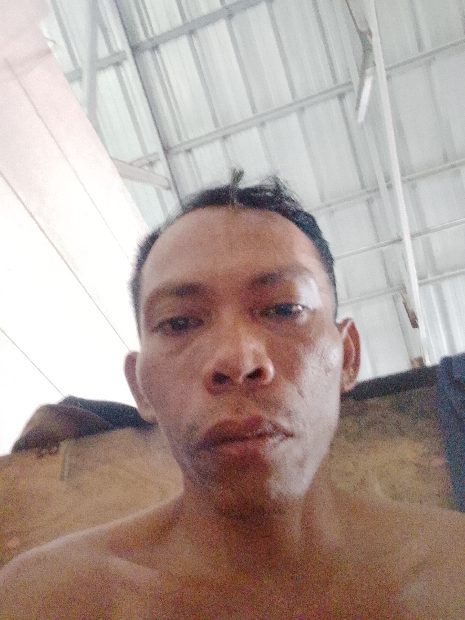 A shirtless man is centered in the frame, facing the camera. He has short, dark hair and a neutral expression. The background features a corrugated metal ceiling with visible beams and lighting fixtures, suggesting an indoor space. A partially visible object or structure is present behind him, with a hint of dark fabric to the right. The location is Bukit Ketok, Indonesia.