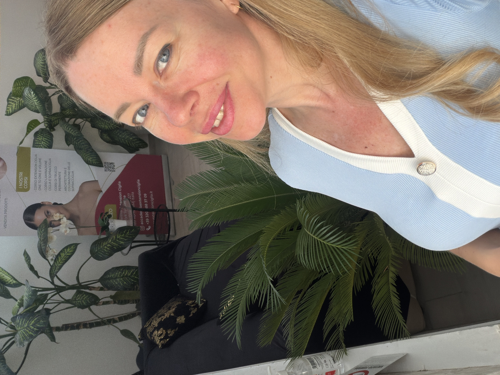A medium shot captures a blonde woman with blue eyes and pink lipstick taking a selfie outdoors, possibly on a balcony, with lush green foliage framing her on the right. Behind her, a dark couch with a patterned pillow is visible, and a large plant with broad leaves stands to her left. A poster with an image of a woman and text in Italian is partially visible in the background, suggesting a beauty salon or training center. The woman's light blue top with white trim and a decorative button are prominent. The lighting suggests it is daytime. The scene does not provide specific clues to pinpoint a location within Rome, Italy.