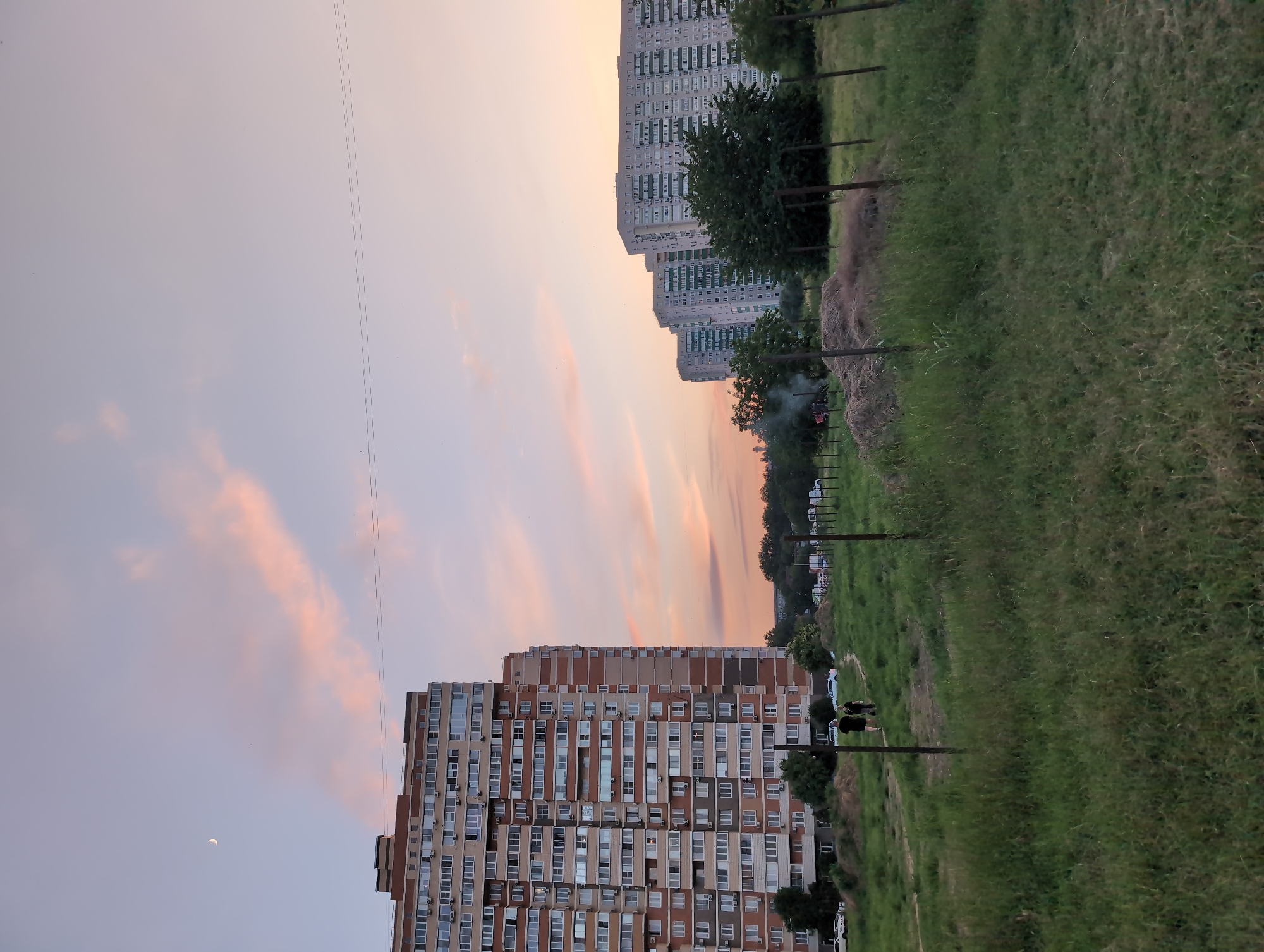 A dusk scene in Krasnodar, Russia, features a row of multi-story apartment buildings. A crescent moon is visible in the pale pink and blue sky, with wisps of clouds tinged with sunset colors. In the foreground, a grassy area is bordered by a wire fence. Two people are walking away from the camera on a dirt path through the grass. Behind them, several other apartment buildings, a line of trees, and a pile of hay are visible. The overall impression is a quiet, residential evening.