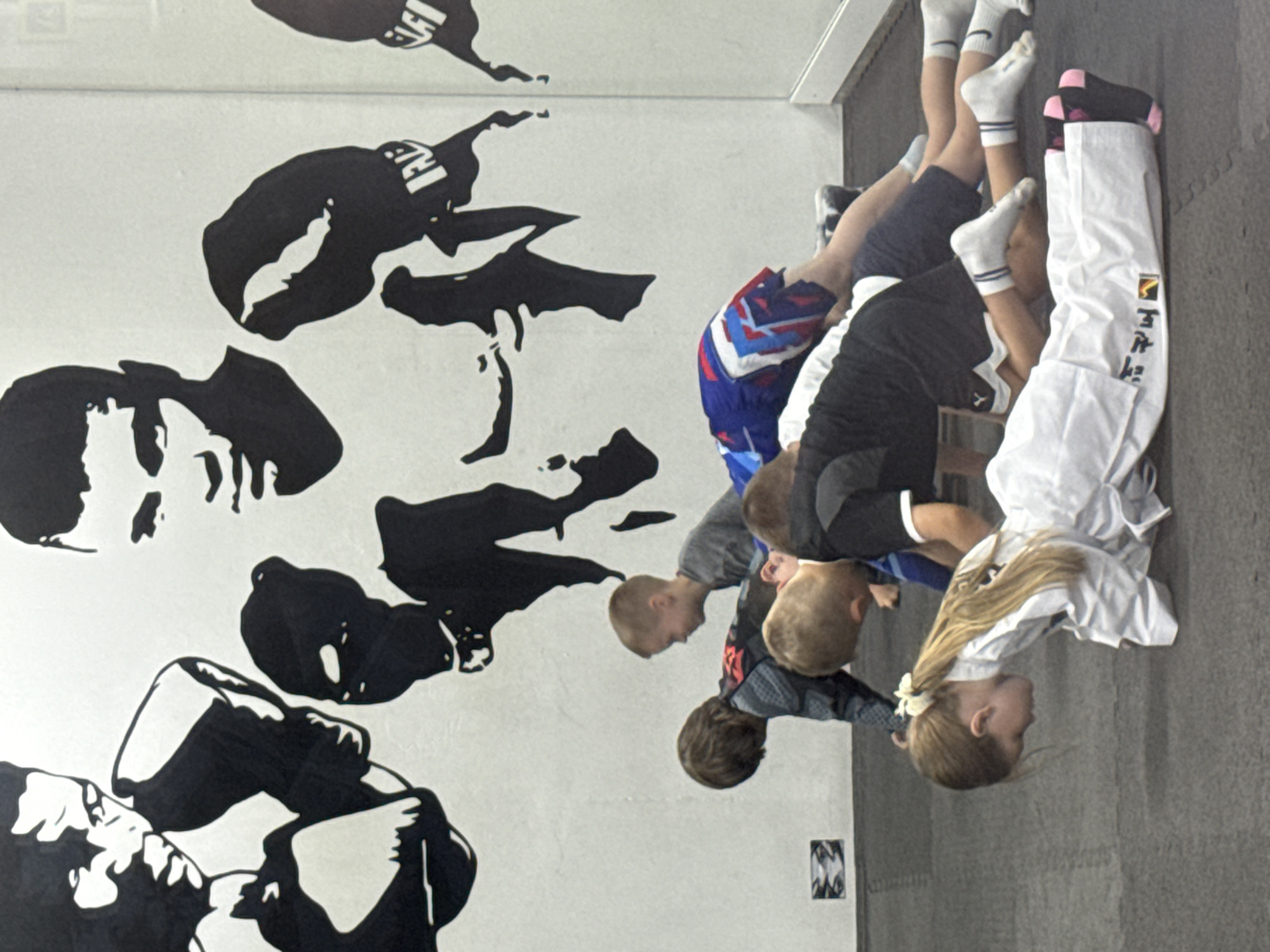 In a room with a black and white mural of a boxer on the wall, several children are performing exercises on a gray mat. The mural features a close-up of two boxers' faces and fists. A young girl in a white martial arts uniform is in the foreground, performing a plank. Behind her, other children are also in plank or push-up positions, wearing athletic clothing. The room appears to be a martial arts or fitness studio. The presence of a prominent mural of a boxer suggests the establishment may be a boxing gym or a martial arts dojo. The overall scene depicts a group of children engaged in physical training.