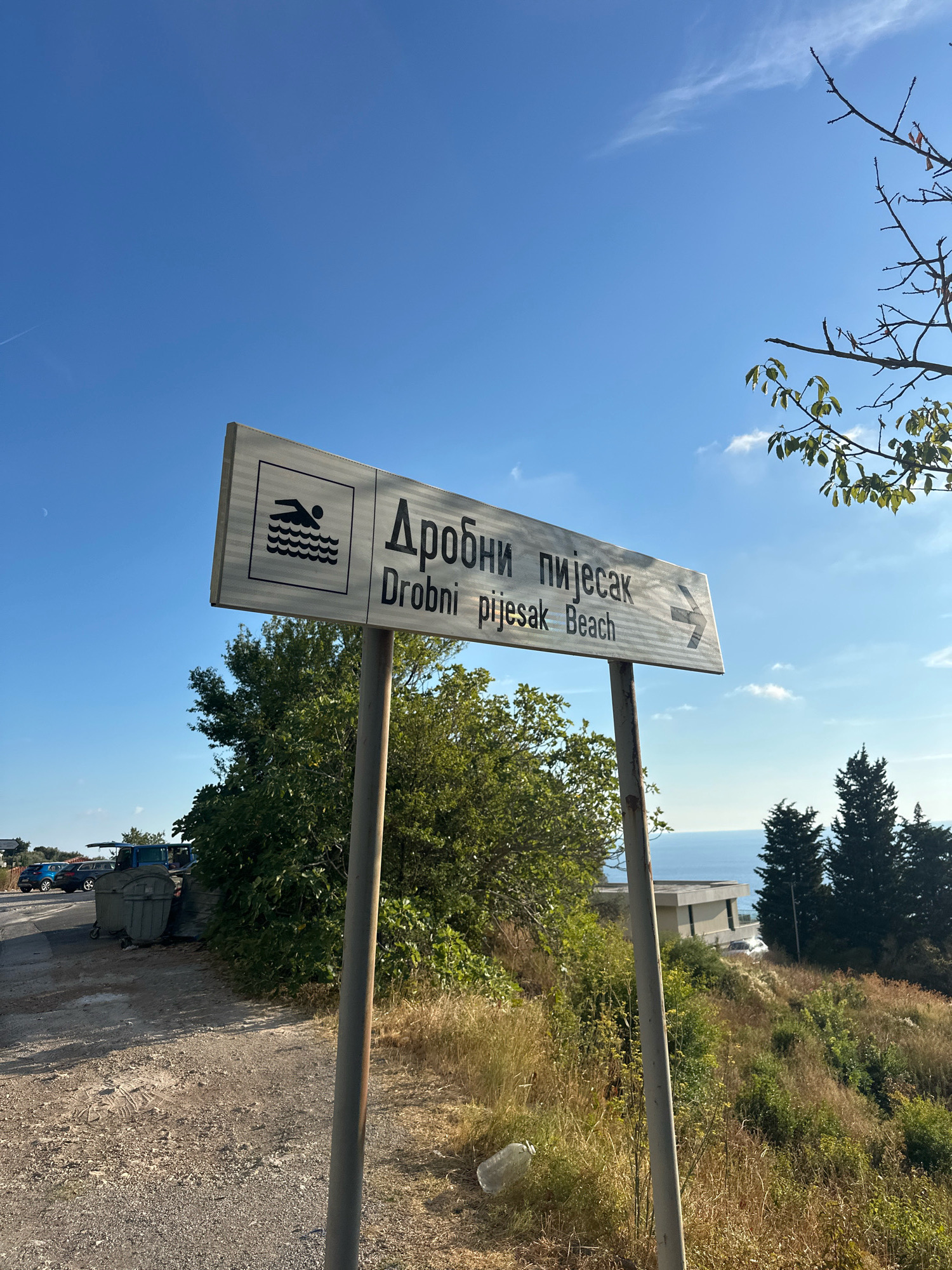 A white signpost stands on a gravel path, indicating directions to "Дробни пијесак" and "Drobni pijesak Beach" with an arrow pointing to the right. The sign features a symbol of a person swimming in waves. To the left of the signpost, a line of parked cars and large green trash bins are visible. Lush green trees and dry grass cover the surrounding hillside. In the distance, a modern building with a flat roof overlooks the blue sea under a clear sky with a few wispy clouds. The scene appears to be a coastal area in Drobnići, Montenegro.