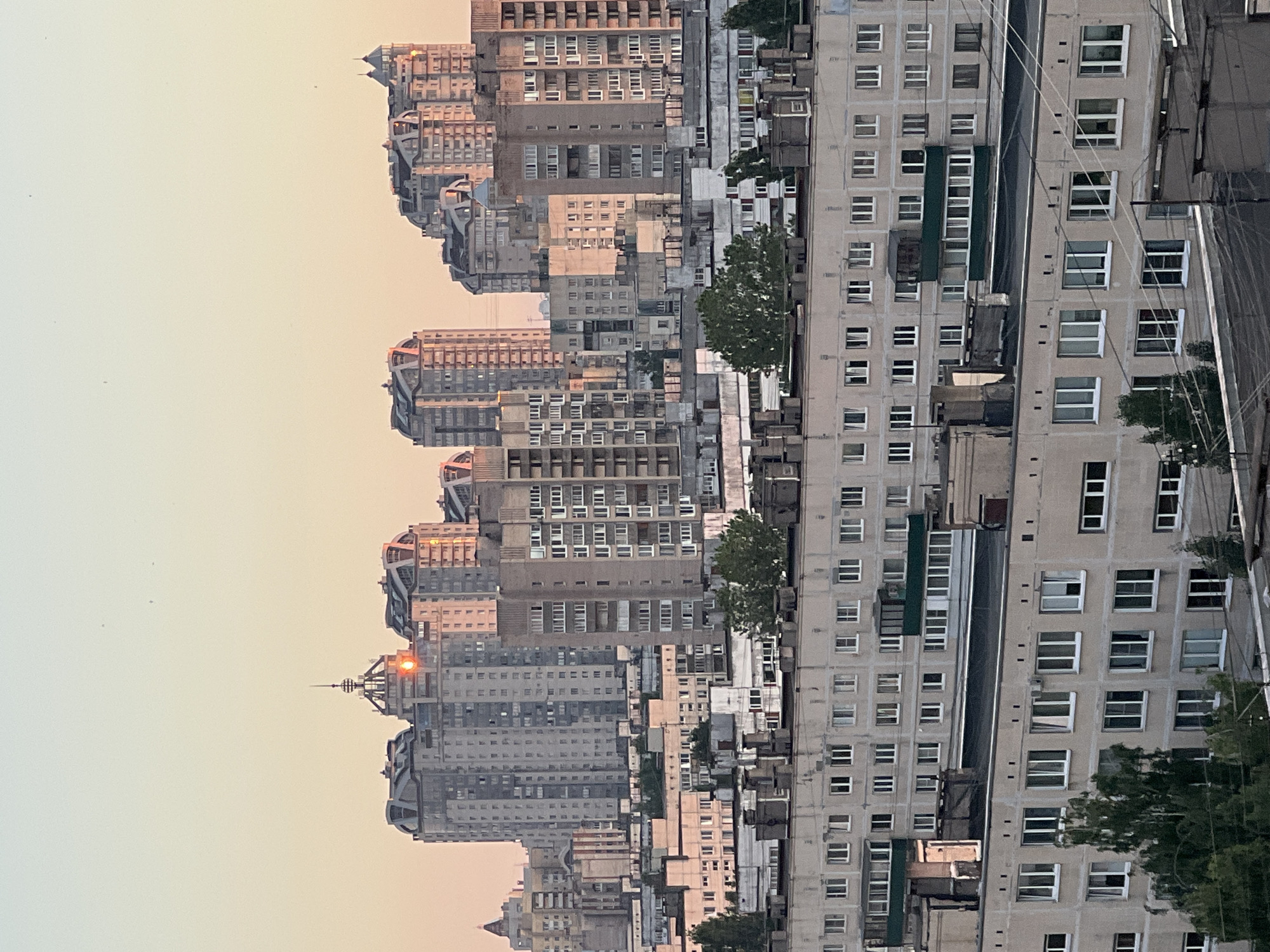 The image depicts an urban skyline at dusk or dawn, characterized by a soft, pastel-colored sky. The scene is dominated by numerous apartment buildings of varying heights and architectural styles, suggesting a densely populated residential area. The buildings in the foreground are lower, featuring a uniform facade with multiple windows. As the eye moves towards the background, taller, more modern structures emerge, some with distinctive curved roofs and decorative elements. The sun is visible as a bright, reddish-orange orb, partially obscured by one of the taller buildings, casting a warm glow on its facade and reflecting in the windows of surrounding structures. This suggests that the photograph was taken from an elevated viewpoint looking westward, capturing the last or first rays of sunlight. The overall impression is one of a vast residential complex bathed in the gentle light of a setting or rising sun. The city of Sinyavino, Russia, is a town in the Kirovsky District of Leningrad Oblast, located on the Neva River.
