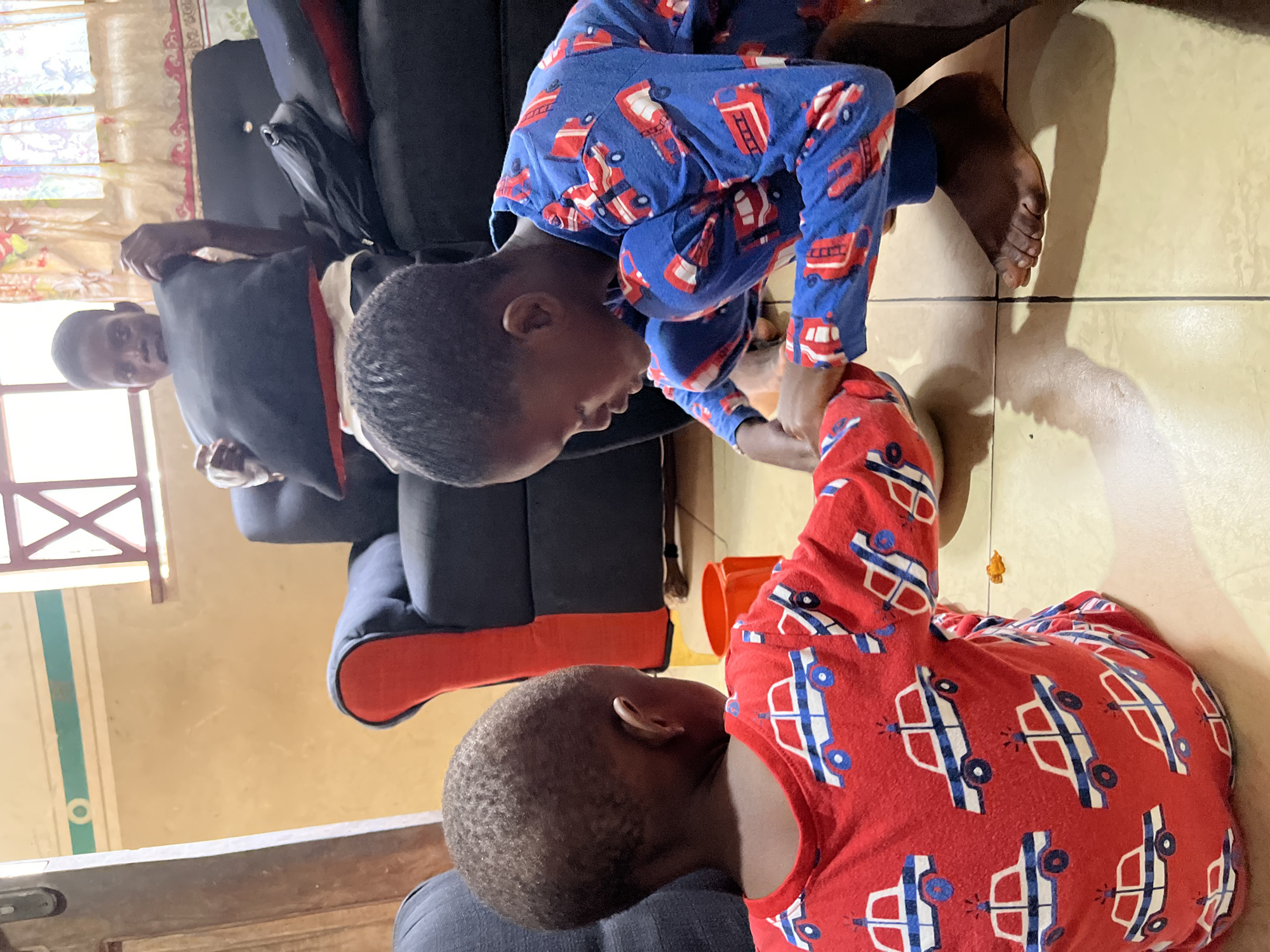 Two children, both in pajamas patterned with cars, are on the floor. One is in red, and the other is in blue. An adult is sitting on a blue couch with red accents in the background. The location appears to be a room with tile flooring and light-colored walls in Badagry, Nigeria.