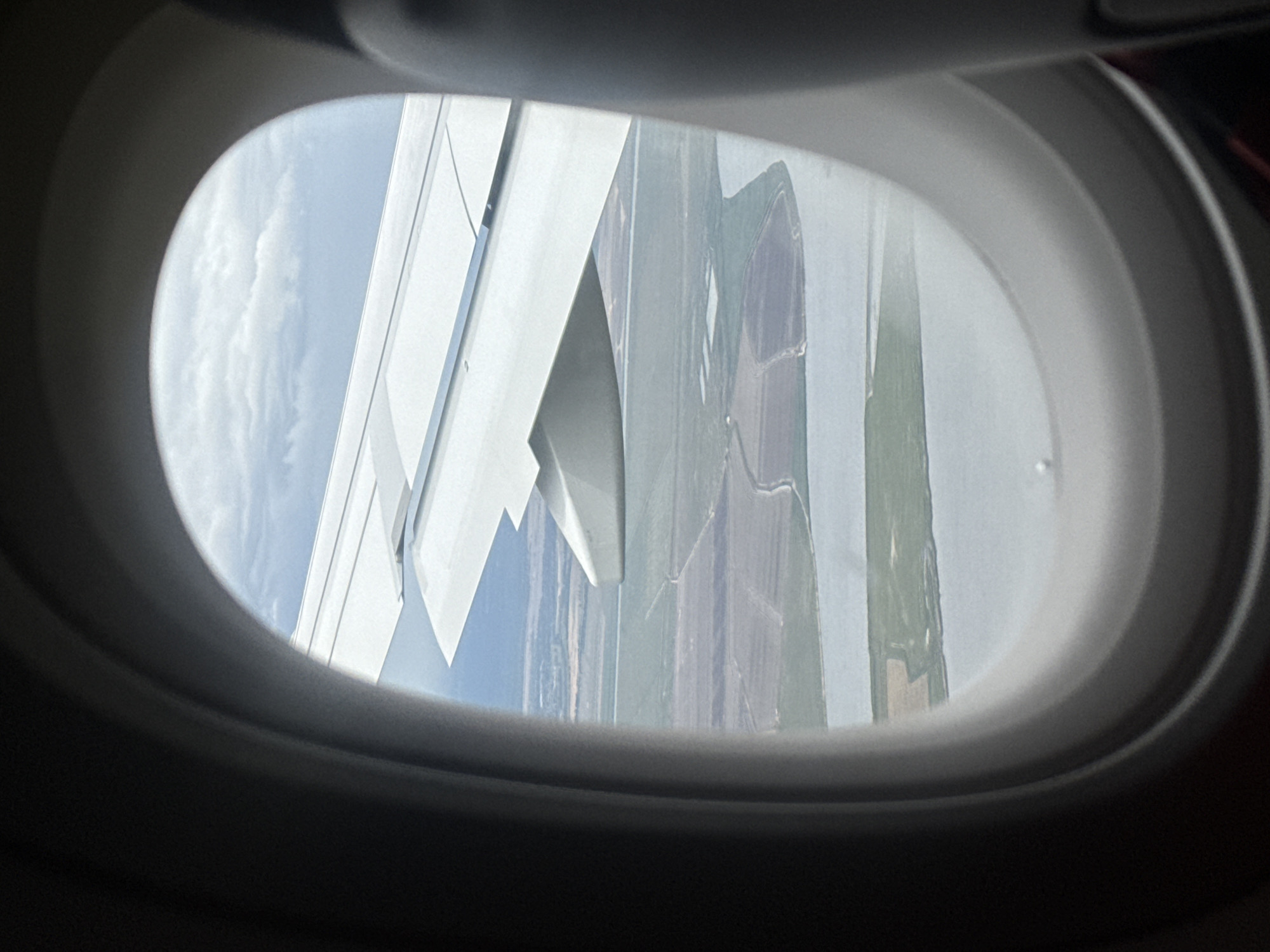 The view from an airplane window reveals a wing and a landscape below. The landscape consists of fields and a river. It's difficult to determine the exact location, the image does not show the location of Montijo, Portugal. No people, subjects, objects, actions, or interactions are visible in the image. The environment is the interior of the airplane and the landscape seen through the window.