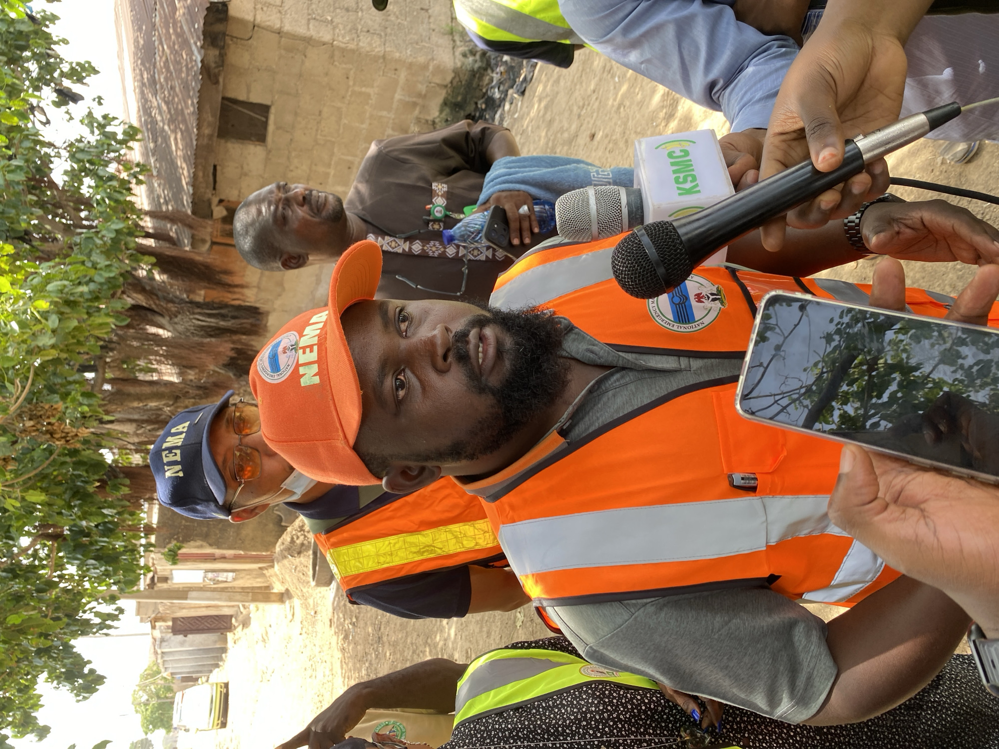 In Sardauna, Nigeria, a man with a beard, wearing an orange baseball cap and a matching safety vest with "NEMA" logos, is the subject of a press interaction. Two microphones are held up to him, one with a "KSMC" logo. An older man is in the background. A second man wearing a baseball cap with "NEMA" is also in the shot. A third person is holding up a mobile phone. A building is partially visible in the background.