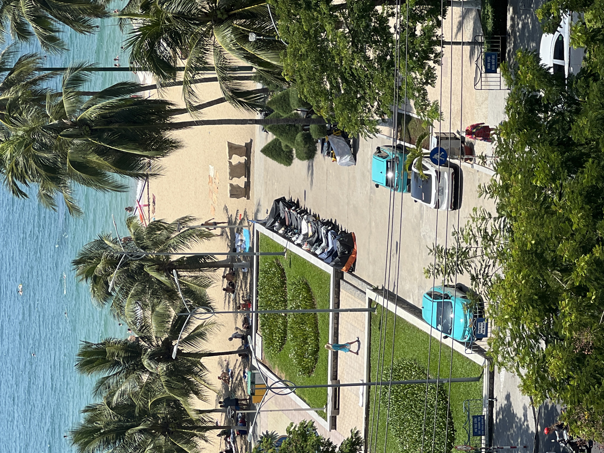 This is an aerial, high-angle shot capturing a lively scene along a beach in Nha Trang, Vietnam, likely in the tourist district. The turquoise ocean, dotted with swimmers and floaters, dominates the upper portion of the image. Palm trees line a sandy beach where several people are relaxing on towels or walking. In the middle ground, a well-maintained green space with manicured bushes separates the beach area from a paved walkway and a parking lot.
Several motorcycles are parked in a line along the right side of the walkway, some covered with tarps. A turquoise car is parked in the lot, and another white car is further down the road. A person in a blue dress is walking on the paved walkway, and a woman in a red outfit is seen walking near a sign in the lower right quadrant. The foreground features lush green trees, suggesting the viewer is looking down from a higher vantage point, possibly a building. The overall impression is one of a sunny, active day at a tropical beach resort.