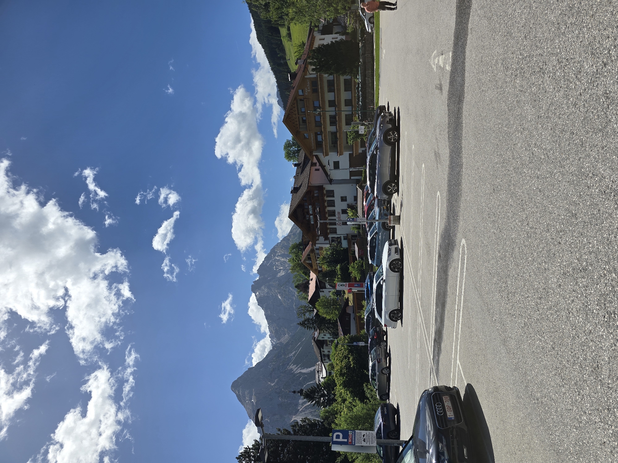 The image shows a parking lot in Lermoos, Austria, with a backdrop of the Zugspitze mountain range. In the foreground, several cars are parked, including a black Audi with a license plate that reads "1-GDP-708". Beyond the parking lot, there are buildings with traditional Austrian architecture, some of which appear to be hotels or guesthouses, with signage like "Haus Lechner". The sky is blue with scattered white clouds, and a green, tree-covered hillside rises behind the buildings. A lone person in an orange shirt is walking away from the camera on the right side of the frame. A streetlamp and a parking sign are visible on the left side of the parking lot.