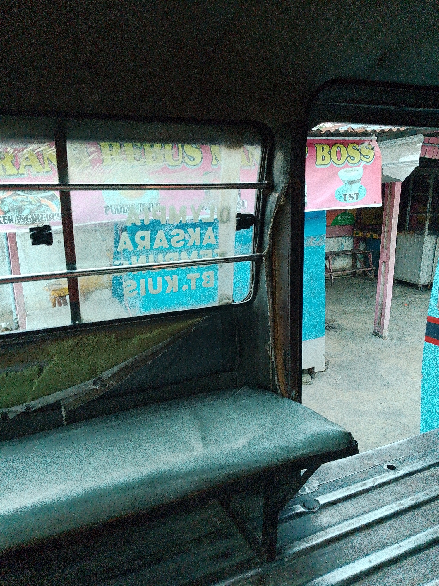 The image shows the interior of what appears to be a public transport vehicle, possibly a bemo or a similar local transportation mode, in Bandar Khalipah, Indonesia. The perspective is from inside the vehicle looking out.
In the foreground, a worn, dark green upholstered bench seat is visible, with a metal frame supporting it. The seat is slightly torn and shows signs of age. Behind the seat, a ribbed metal surface, likely the floor or cargo area of the vehicle, is visible.
Through the windows, there are several signs and elements of the outdoor environment. On the left, a window shows a sign with text, partially obscured and possibly in Indonesian. Some of the visible text includes "KERANG REBUS" and "PUDING". On the right, through another opening or a broken window section, a prominent pink banner with the word "BOSS" in yellow lettering is displayed. Below "BOSS", a stylized drawing of a drink or food item is present, and beneath that, "TST" is written. Further to the right, a blue structure and a wooden bench can be seen, as well as some metal cabinets or carts. The ground appears to be a paved or concrete surface.
The overall lighting suggests it is daytime, and the scene depicts a common urban or semi-urban setting in Indonesia, characterized by local businesses and transportation.