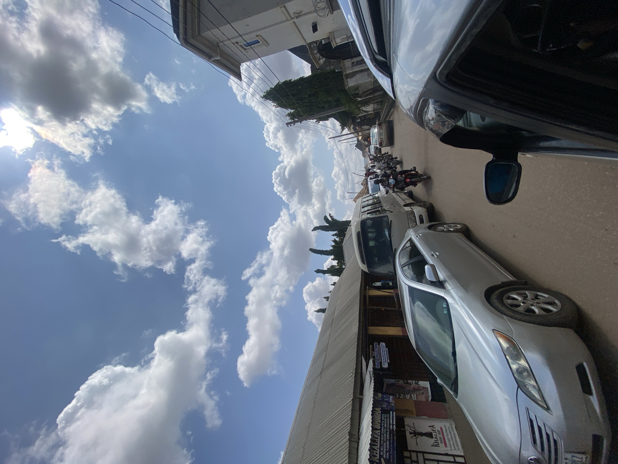 The image captures a street scene in Ado, Nigeria, under a partly cloudy sky. In the foreground, the side and partial interior of a silver car are visible, suggesting the photograph is taken from within a vehicle. The street is congested with traffic, featuring a silver Toyota Camry in the left lane, a white mini-bus, and several motorcycles. A line of vehicles and motorbikes is moving down the road, indicating moderate traffic. On the left side of the street, there are commercial establishments with corrugated metal roofs and signage, one of which reads "KHALIFA SHISHA". The buildings on the right appear to be residential homes, some with modern architectural styles. The overall environment depicts a typical urban street during daylight, with elements of commerce and transportation.