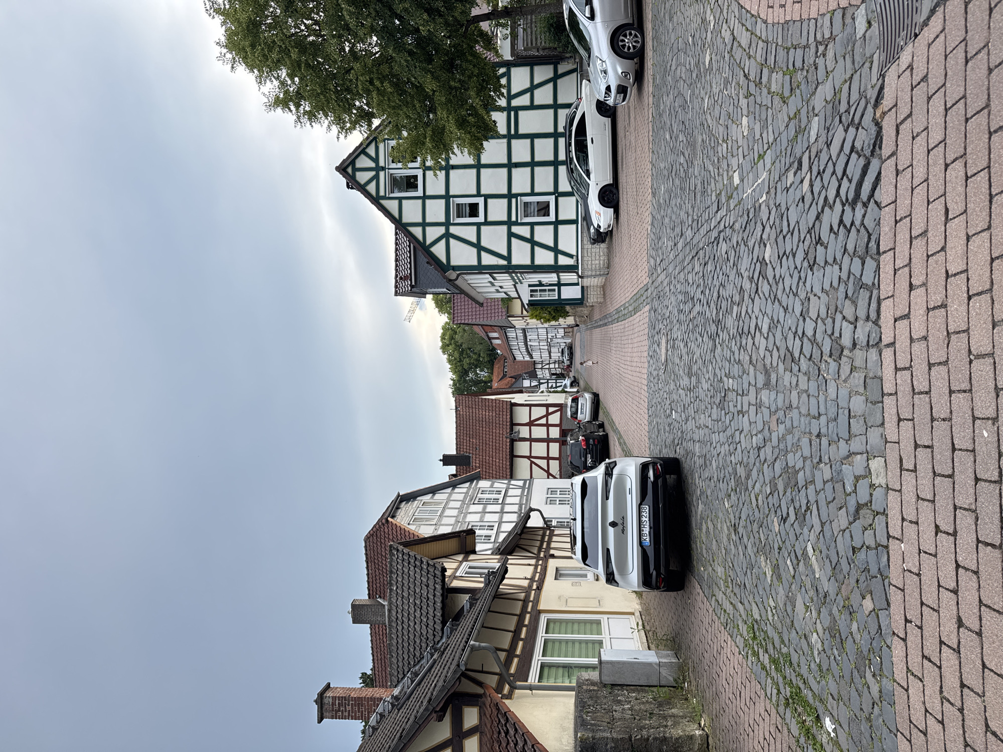 The image depicts a narrow cobblestone street in a European town, characterized by traditional half-timbered houses. The sky is overcast. On the left side of the street, a white SUV is parked, facing away from the camera. Further down the street, several other cars are parked. The architecture of the buildings features dark wooden beams against white plaster, with red tiled roofs. Trees line the right side of the street. A small child is visible in the distance, walking on the left side of the street. The cobblestone pavement is a mix of grey and reddish-brown stones, with some grass growing in the gaps.