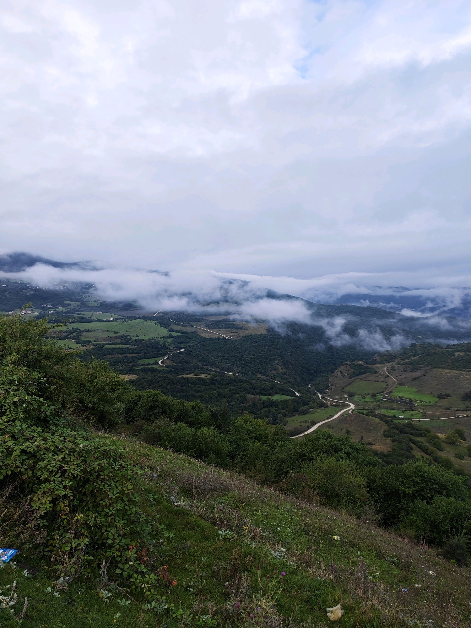 A drone shot captures a panoramic view of a valley shrouded in clouds on a cloudy day. In the foreground, a steep, grassy slope is covered with wild bushes and vegetation, with a scattering of litter visible near the bottom left. The midground reveals a winding road cutting through lush green hills and dense forests. The background is dominated by a range of mountains partially obscured by swirling clouds, suggesting a misty or foggy atmosphere. Scattered houses and fields are visible on the slopes, indicating a rural settlement within the valley. The overall scene depicts a serene and somewhat mysterious natural landscape, likely in the mountainous region of Цухдыг, Russia.