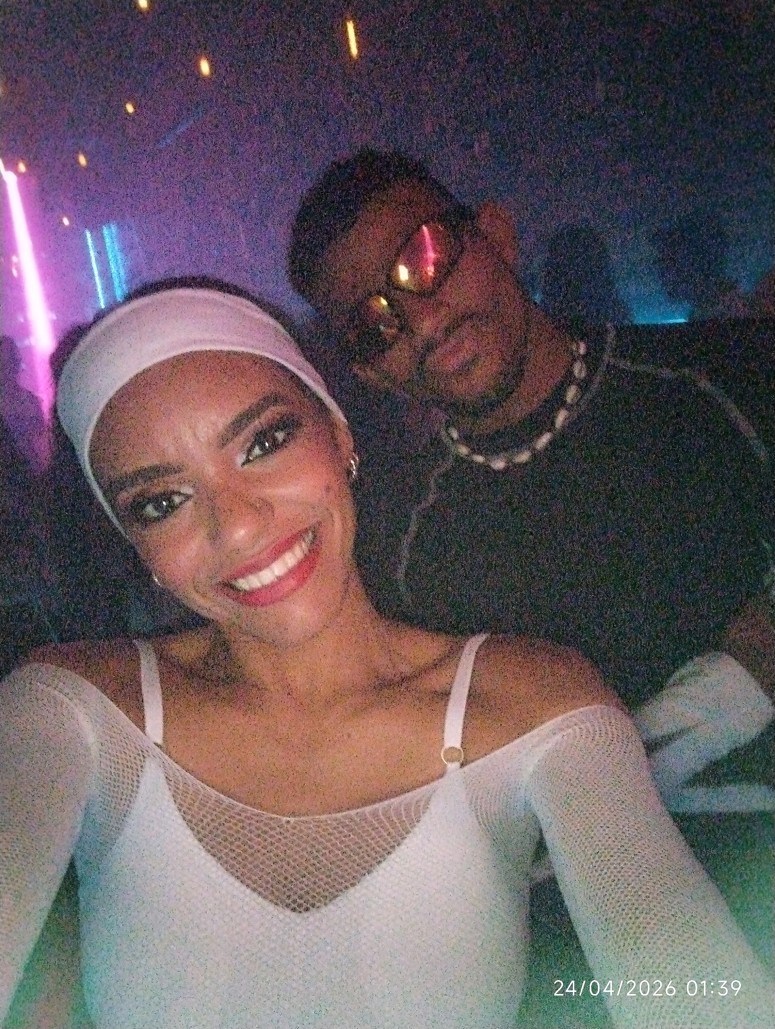 A photograph shows two individuals in what appears to be a nightclub or similar dimly lit venue. A woman is in the foreground, wearing a white headband, a white mesh top with spaghetti straps, and matching mesh long sleeves. She has visible makeup and is smiling at the camera. Behind her and slightly to the right is a man wearing sunglasses, a black shirt, and a beaded necklace. The lighting in the background is colorful, with pink and blue hues. The date "24/04/2026 01:39" is visible in the lower right corner. The location is unspecified.