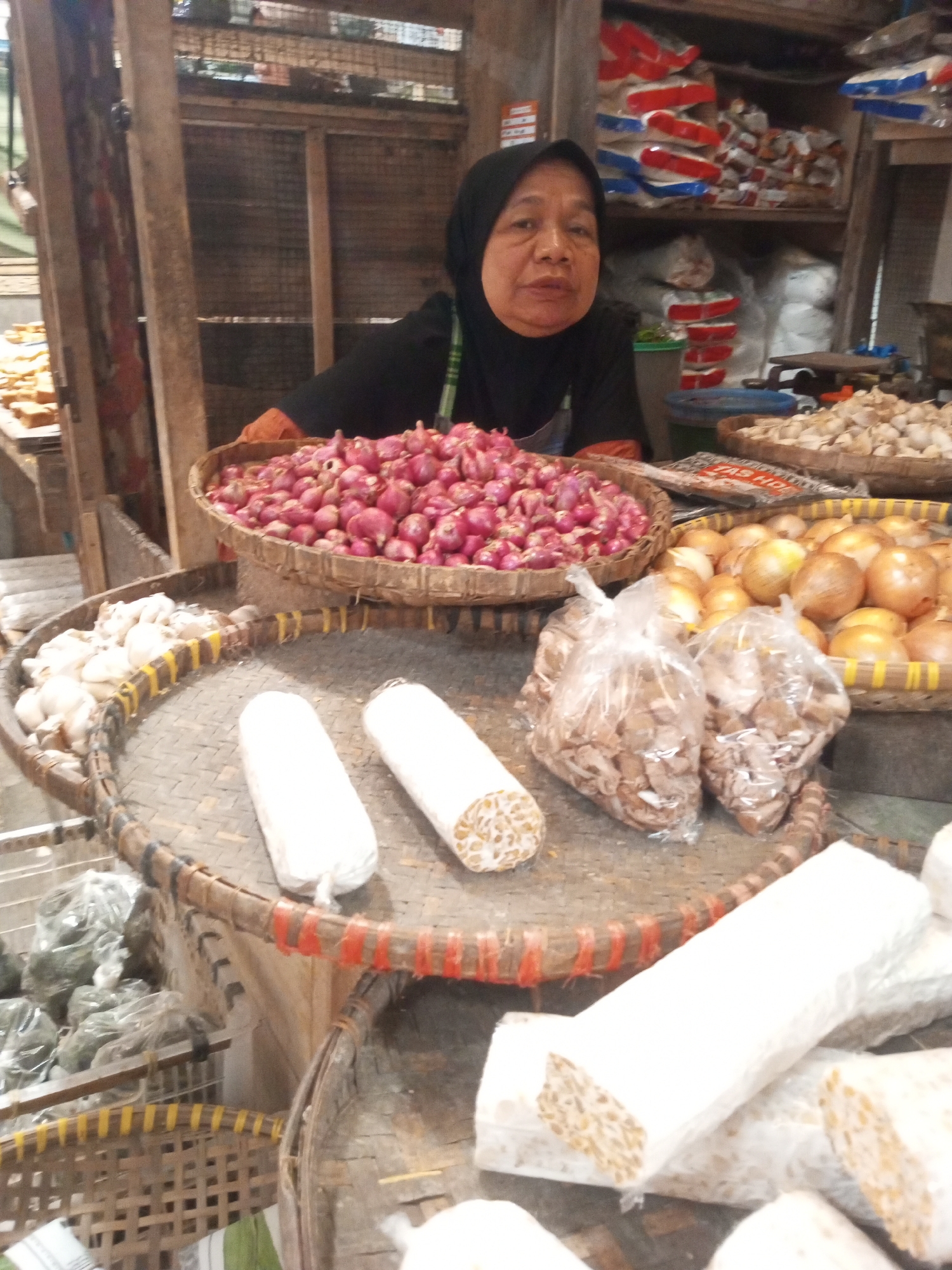 In Kalibeber, Indonesia, an outdoor market stall displays a variety of produce. A woman wearing a black hijab and black top stands behind a woven basket filled with red onions. In front of her, on a larger woven circular tray, are two white-wrapped packages of tempeh, and to the right, a basket overflows with yellow onions. Another woven tray to the left holds a pile of garlic. Behind the woman, shelves are stocked with bagged goods, some with red and blue packaging. The stall is constructed of wood.