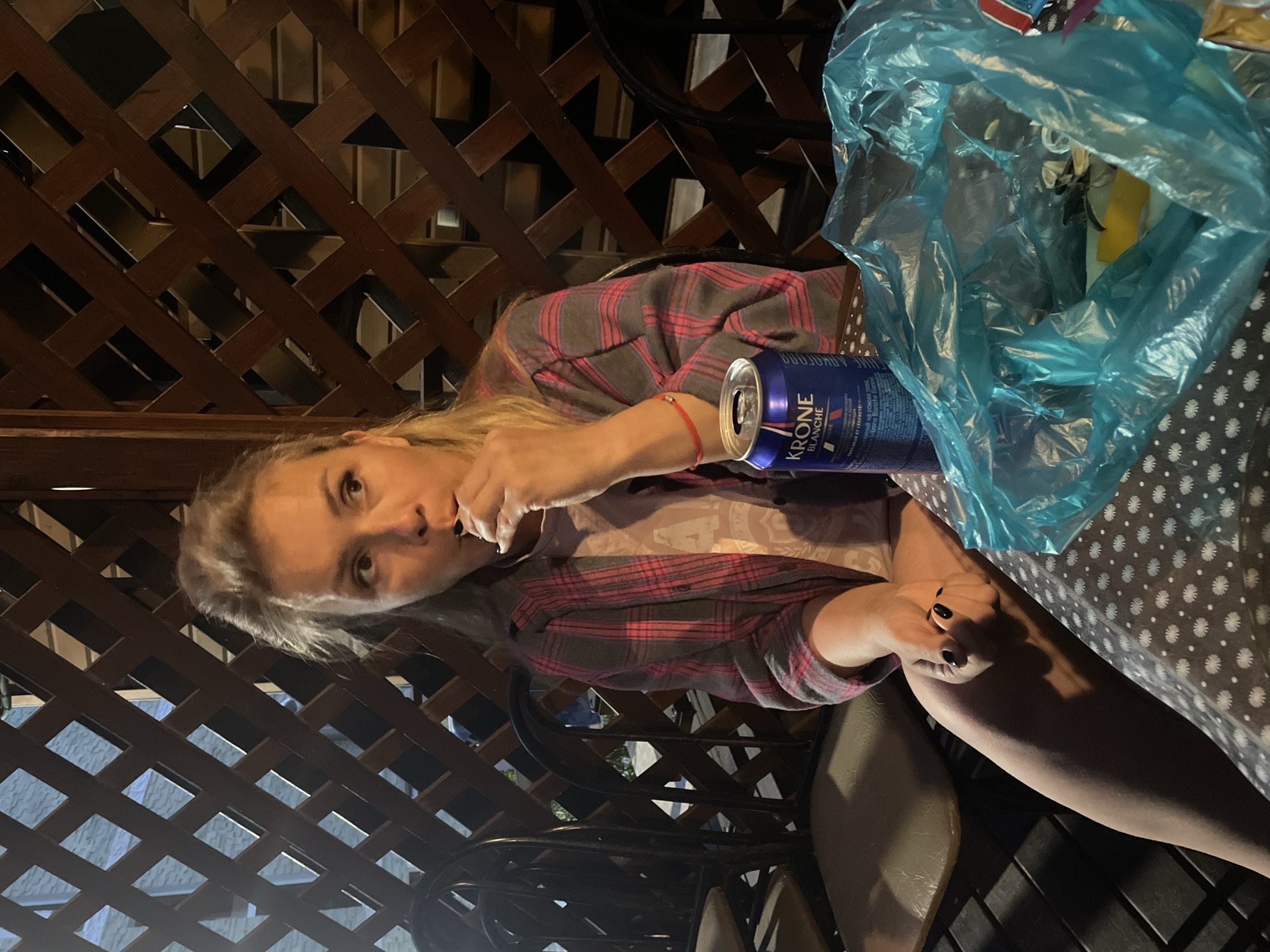 An outdoor, nighttime scene features a woman with blonde hair sitting at a table. She is wearing a pink and grey plaid shirt over a lighter-colored shirt and has a red bracelet on her left wrist. The woman is bringing a piece of food to her mouth with her right hand. Her nails are painted black. In front of her on the table is a blue can of Krone Blanche beer and a bright blue plastic bag filled with what appears to be trash. The table is covered with a tablecloth patterned with white flowers on a dark background. The woman is seated at a wooden lattice structure that provides a backdrop, and chairs are visible to her left. The lighting is dim, suggesting an ambient outdoor setting.