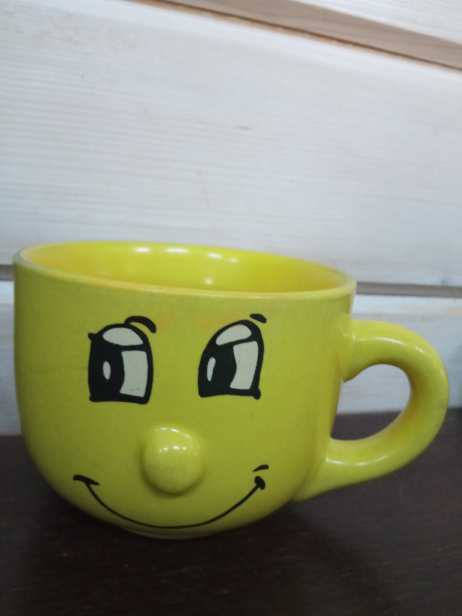 A bright yellow ceramic mug with a cartoonish smiley face design sits on a dark brown surface. The mug features two large black and white eyes, a small raised nose, and a curved black line for a smile. The mug has a curved handle on its right side. In the background, a light-colored wooden wall with horizontal planks is visible. The scene is indoors, and the lighting appears to be neutral. The location is Finskiy Briz, Russia.