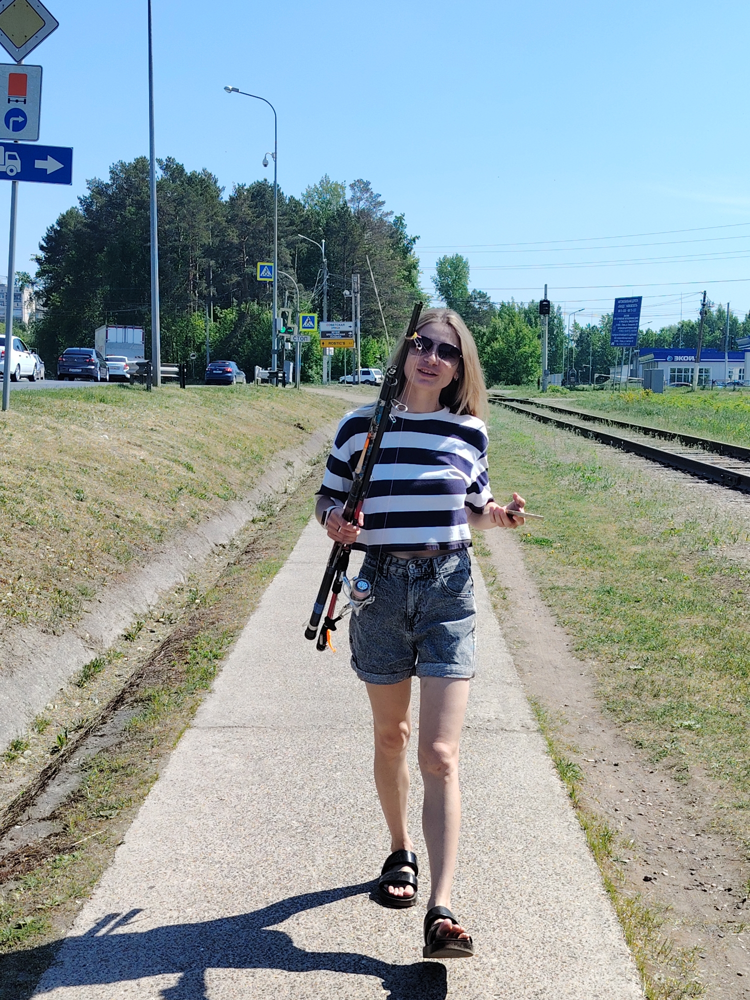 A woman walks on a concrete path next to a set of railroad tracks, holding fishing rods in her left hand and a small wooden object in her right. She wears sunglasses, a striped shirt, denim shorts, and sandals. The path is flanked by grass and a drainage ditch on the left and the railroad tracks on the right. In the background, there is a road with traffic signs, trees, and buildings. A blue sign with white lettering is visible beyond the railroad tracks. The location appears to be Chaikovsky, Russia, near the "ЭКОЙЛ" gas station.