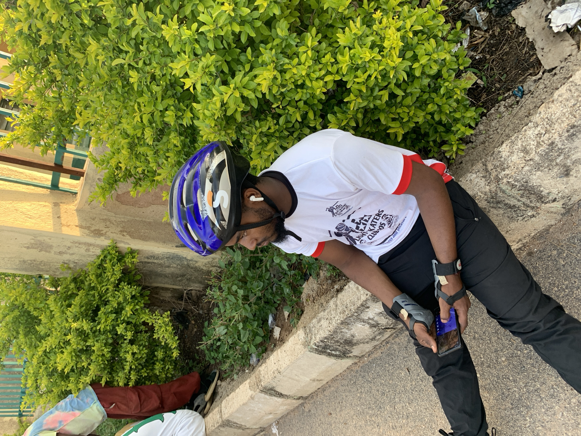 An outdoor scene depicts a dark-skinned male seated on a concrete curb, adjacent to an asphalt road. He is wearing a blue, black, and white cycling helmet with an orange "S" logo, a white wireless earbud in his right ear, a white short-sleeved t-shirt with a black collar and red sleeve trim, black trousers, and grey and black wrist guards on both wrists. The t-shirt bears the text "LIGHT SKATERS CLUB JOS" and imagery of inline skates. He holds a black smartphone horizontally with both hands, looking at its screen.
Behind the man, a dense, vibrant green bush occupies the space between the curb and a light brown/beige concrete wall or building structure. Further in the background, above the bush, a green metallic fence or gate is visible. The ground behind the curb consists of dirt with scattered green foliage and some small pieces of litter. The concrete curb itself shows signs of wear and discoloration.
To the left and slightly behind the main subject, another individual is partially visible, seated or bent over. This person wears a multi-colored tie-dye t-shirt (light blue, pink, yellow/orange) and dark red/maroon cargo pants. Dark footwear is also visible. The lighting is bright natural sunlight, indicating daytime. The scene presents an urban or semi-urban environment consistent with locations found in cities such as Jos, Nigeria.