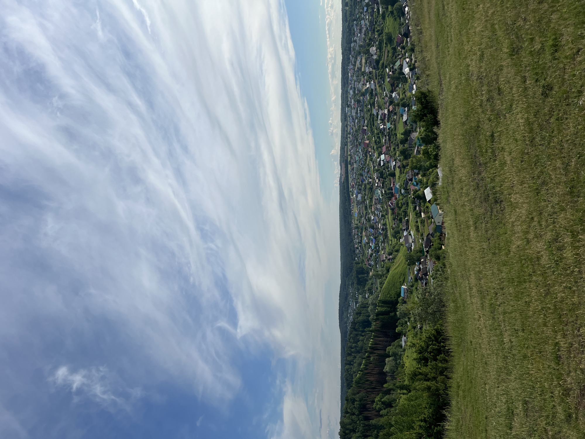 The image captures a wide-angle landscape view, likely taken from an elevated position overlooking a rural or semi-rural area in or near Ольховка, Russia.
The upper portion of the frame is dominated by a clear blue sky, interspersed with long, wispy white clouds, characteristic of cirrus formations, stretching horizontally across the expanse. Below the sky, on the left side of the scene, lies a sprawling settlement. Numerous small houses, each with a distinctively colored roof (ranging from red and blue to green and grey), are visible, nestled amidst verdant trees and patches of green. This indicates a village or small town extending along a distant ridge.
A band of dense green forest forms a natural boundary between the sky/settlement and the foreground, running along the horizon line. The right side and the entire foreground are occupied by a vast, gently sloping field of tall, lush green grass, suggesting a pastoral setting. No specific human activity or identifiable individuals are visible within the frame.
The scene is illuminated by bright daylight, suggesting fair weather during the late morning or afternoon. The overall impression is one of peacefulness and natural beauty, combining elements of human habitation with expansive natural landscapes. There is no visible text in the image.