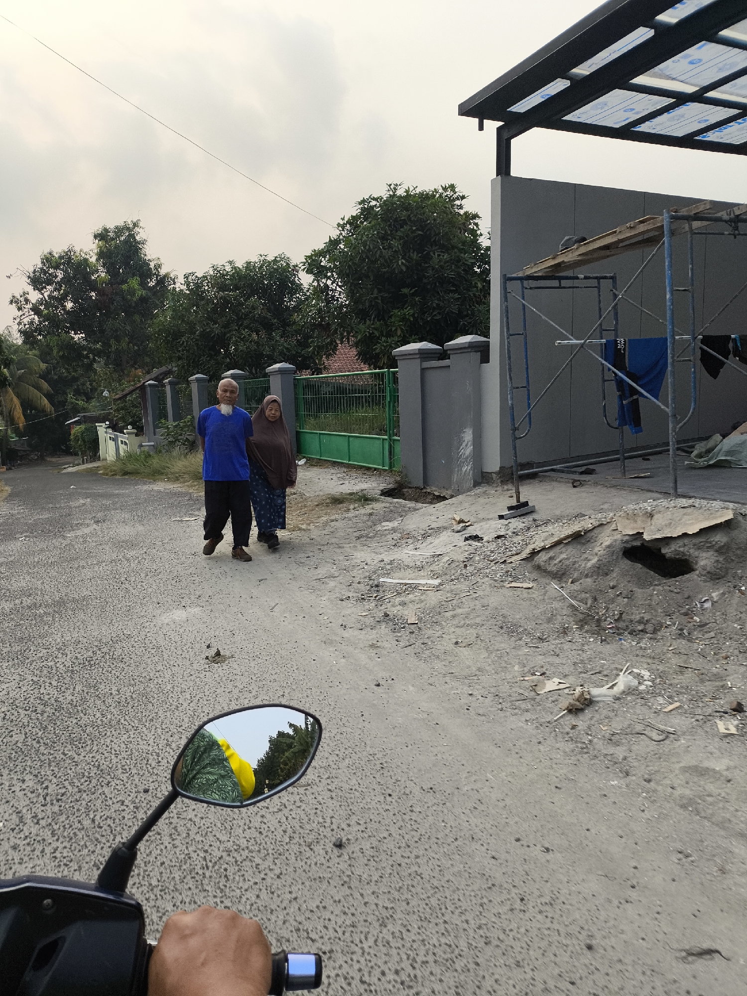 In Cilegon, Indonesia, on an overcast day, two individuals are walking on a paved road in front of a partially constructed building. The building has a dark gray facade and a modern, dark-framed canopy with translucent panels. Blue scaffolding is erected against the wall, with some clothing hanging from it. The foreground of the image is dominated by the road and construction debris, including dirt, rocks, and scattered pieces of wood and plastic. A motorcycle's side mirror is visible in the lower left corner, reflecting the sky and trees, and a hand is on the handlebar. The two pedestrians, an older man with a white beard and a blue shirt and dark pants, and an older woman in a brown hijab and patterned skirt, are walking away from the viewer towards the construction site. Behind them, a green fence and trees line the road, with a house visible in the background. The overall scene depicts a residential area with ongoing construction.