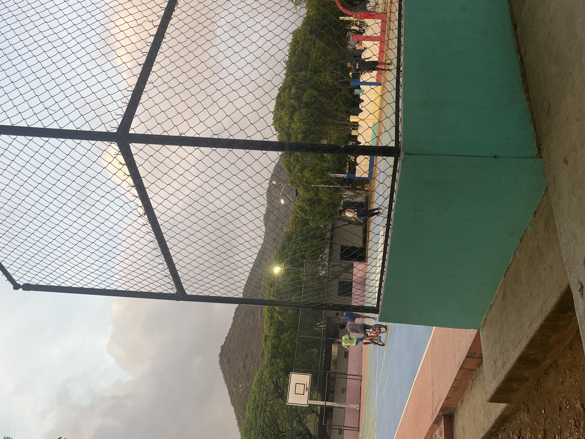 The image shows a fenced outdoor basketball court in Santa Ana, Venezuela. The court is blue and red with a white basketball hoop. Several young people are present, some riding bicycles. In the background, there are trees and a mountain. The sky is cloudy.