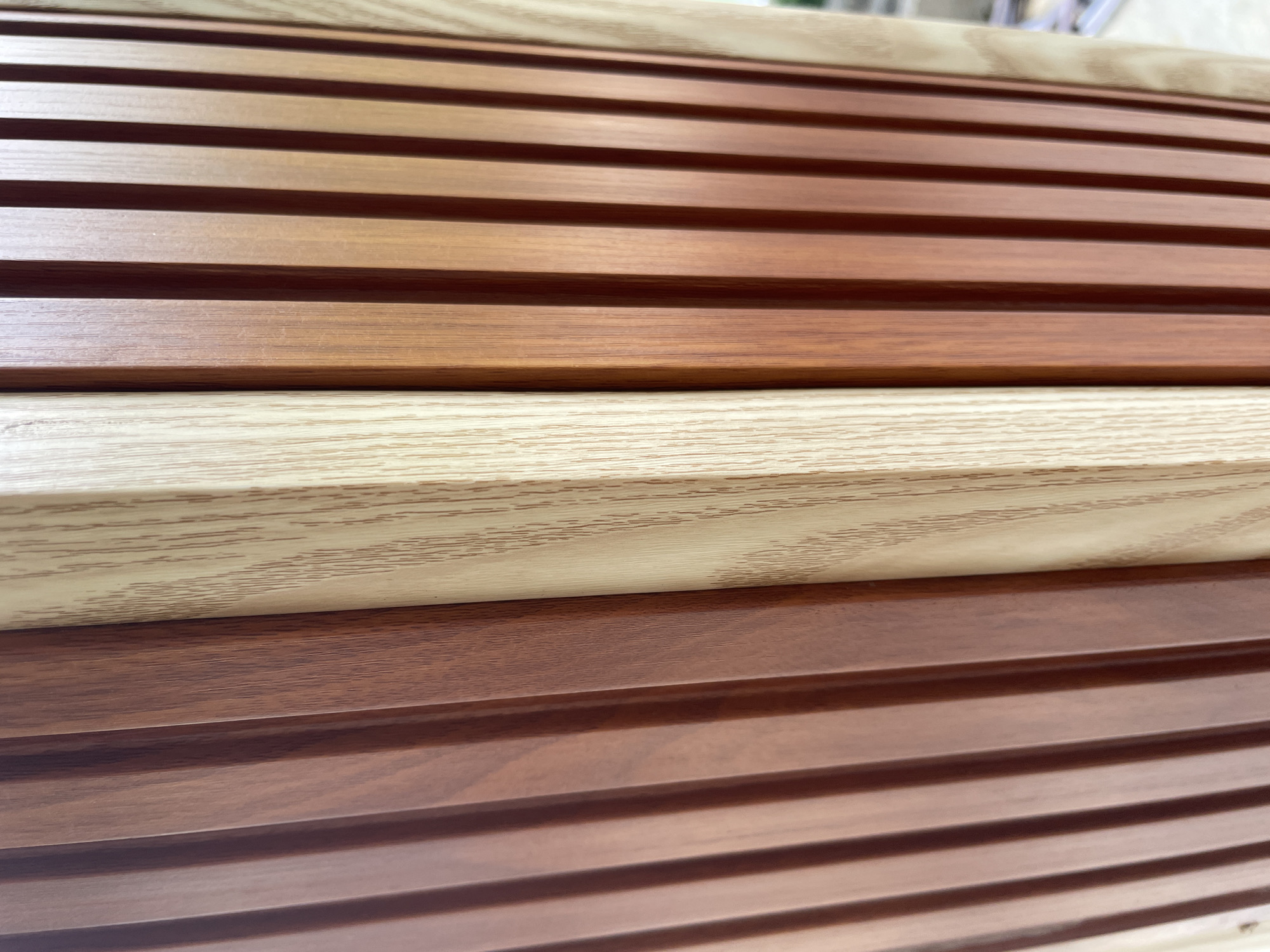 The image shows a close-up, eye-level view of vertical wooden slats. On the left side of the image, there is a section of lighter-colored wood with a visible grain pattern. To the right of this, there are several slats of a darker, reddish-brown wood. The lighting suggests an outdoor environment, as there are shadows and highlights on the wood surfaces, indicating a light source from the upper right. The image is devoid of any people, actions, or interactions. Based on the visual information provided, it is impossible to determine the specific location within Phan Thiet Ward, Vietnam.
