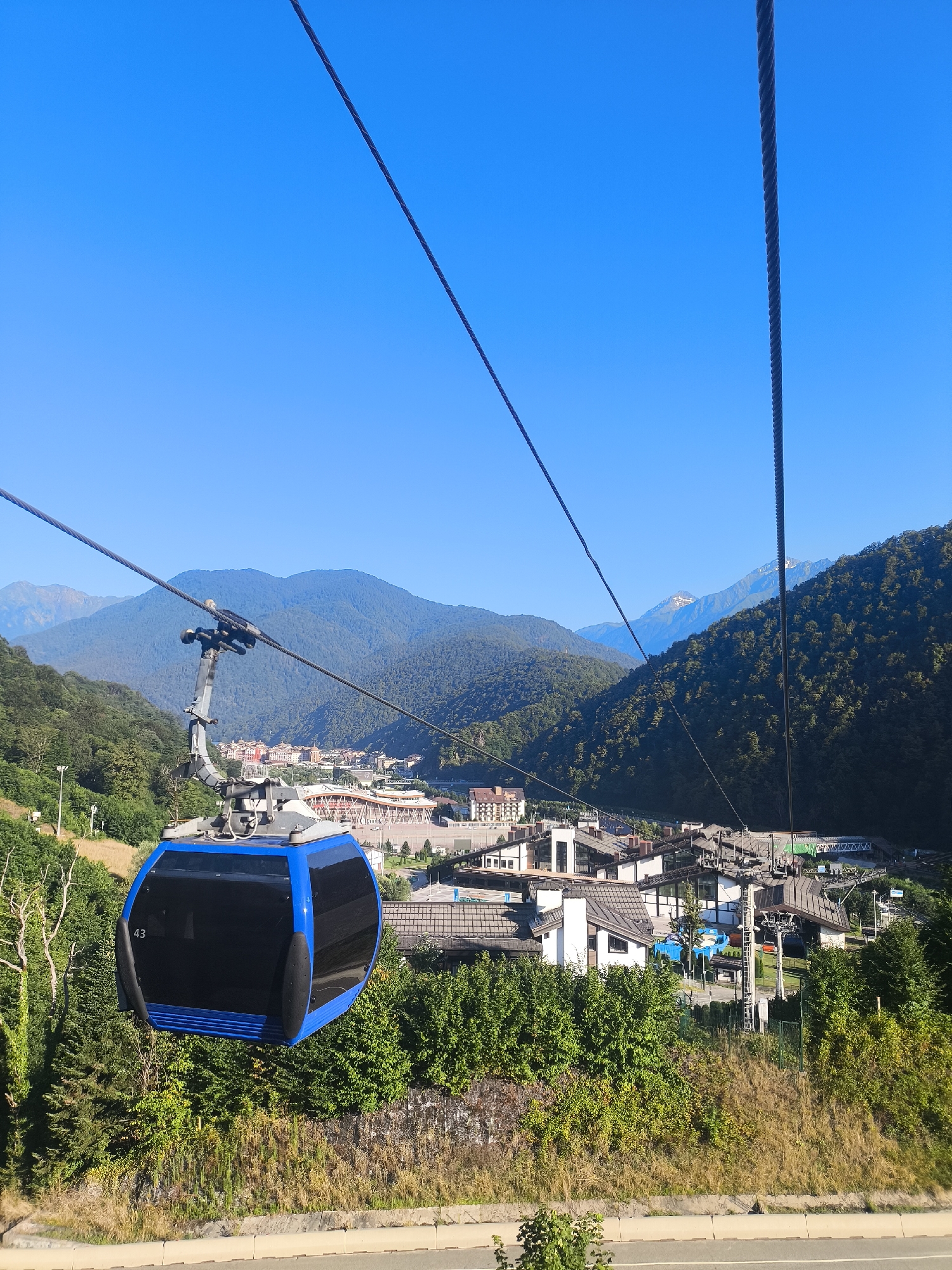 This is a ground-level, eye-level, medium shot of a blue and black gondola on a cable car system, moving towards the viewer. The gondola is in the foreground and is numbered "43" on its left side. The background features a mountainous landscape with a town nestled in a valley below. The town has numerous buildings with light-colored walls and dark, sloped roofs, suggesting a resort or ski village. Several cable car towers and ski lifts are visible throughout the town. Lush green trees and vegetation cover the slopes of the mountains, and the sky is a clear, bright blue. The scene is set in Estosadok, Russia.