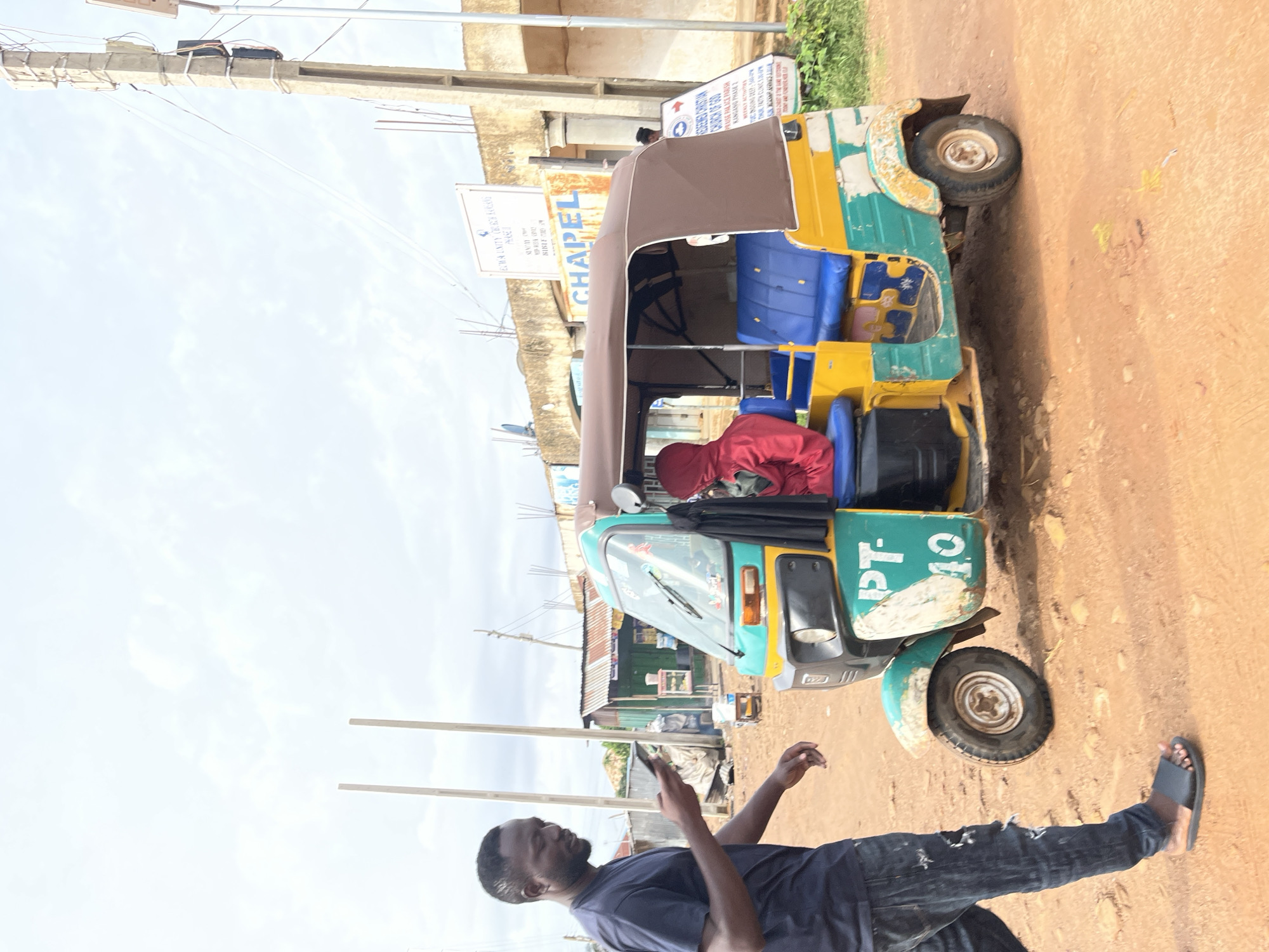 In the city of Jos, Nigeria, a man in a dark blue t-shirt and ripped black jeans stands to the left, looking down at his phone. He is walking next to a colorful auto-rickshaw, which is green, yellow, and white, with blue seats. The auto-rickshaw has "PT-10" written on its side. A person wearing a red hooded jacket is seated inside the auto-rickshaw. In the background, there are buildings, including one with a sign that says "CHAPEL" and another with the text "CHURCH OF GOD". There are also utility poles and wires visible. The ground is covered in reddish-brown dirt, indicating an outdoor, possibly unpaved, area. The sky is cloudy.