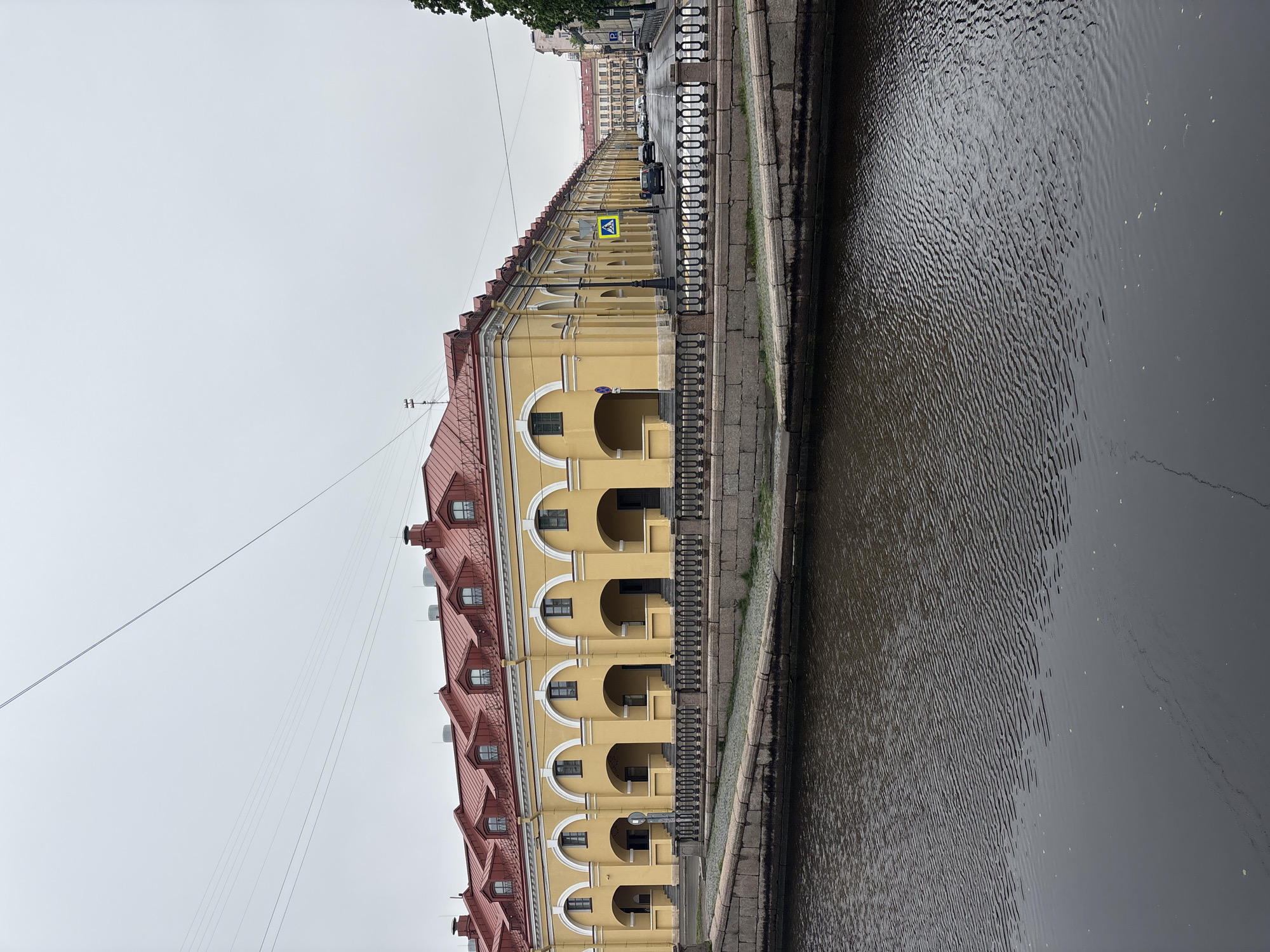 The image depicts a long, yellow building with a red roof, showcasing an arcade structure with arched openings along a riverbank in Saint Petersburg, Russia. The building features multiple stories, visible through the arched openings, and dormer windows in the roof. A stone embankment runs alongside the building, bordering the river, which shows ripples on its surface. A pedestrian crossing sign is visible along the building's facade, suggesting a street. The sky is overcast, providing soft, diffused lighting. Cars and other buildings can be seen in the distance. A metal railing runs along the walkway beside the river. Overhead wires are visible across the sky.