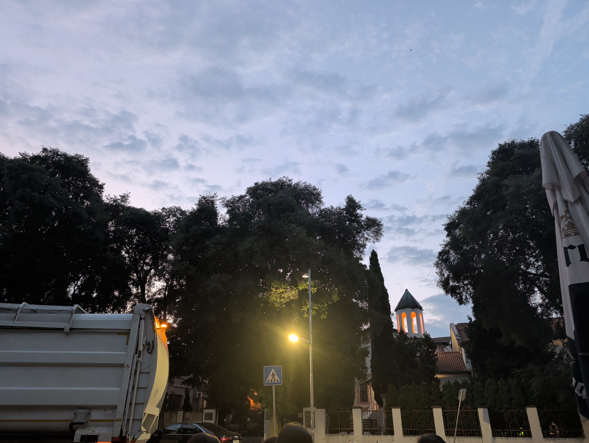 The image depicts a street scene at dusk in Varna, Bulgaria. In the foreground on the left is the side of a white garbage truck. In the midground, a street lamp illuminates a pedestrian crossing sign and the dense foliage of trees. Behind the trees, a building with an illuminated tower, possibly a church, is visible. The sky above is cloudy and a pale blue, suggesting the twilight hour.