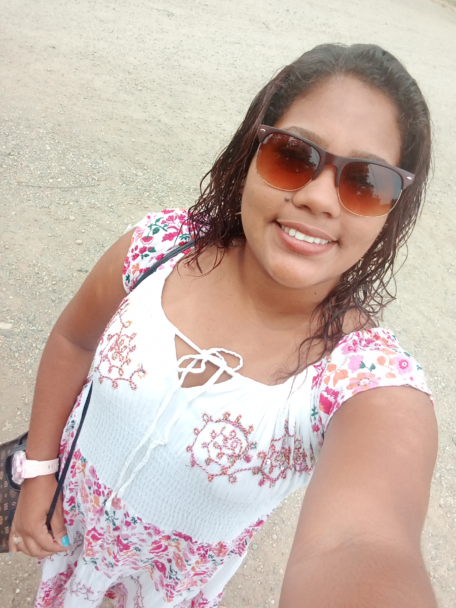 A woman is posing for a selfie outdoors in Parroquia Ocumare, Venezuela. She is wearing sunglasses, a white dress with floral patterns, and a pink watch. A purse is slung over her shoulder. The background is a gravelly or dirt surface.
