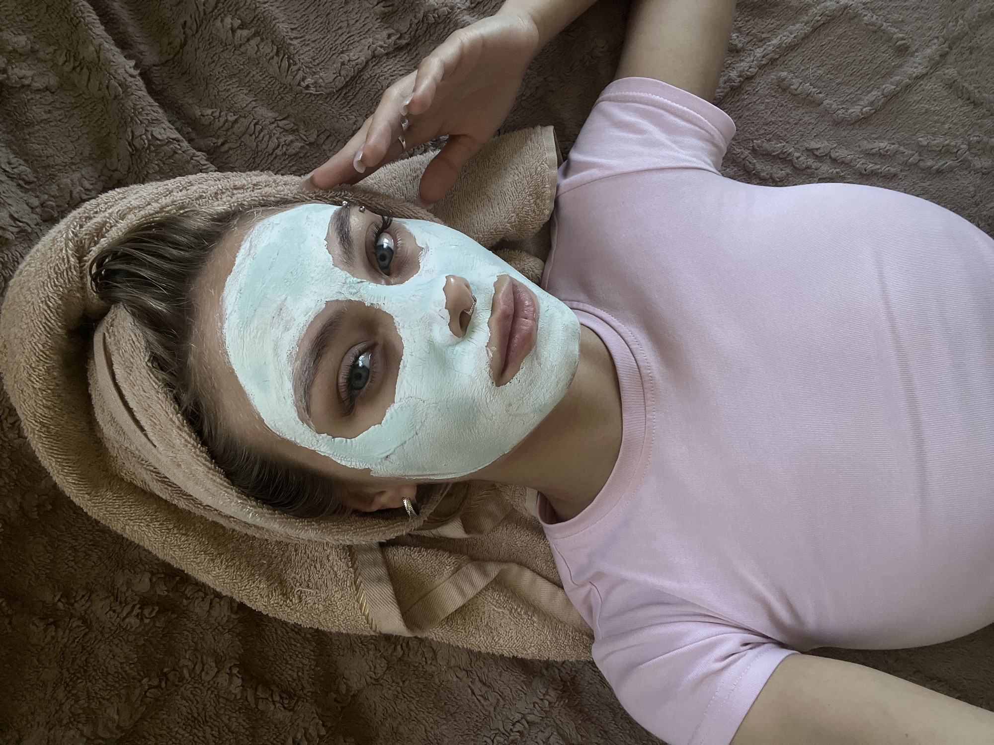 A young woman with blonde hair pulled back by a towel is lying down, applying a light blue facial mask. She is wearing a pink t-shirt and has a nose ring and an earring. She is looking up at the camera, with one hand resting near her face. The background is a brown, textured surface, possibly a blanket or bedspread. The image does not contain any information about the location in the city of Фоки, Russia.