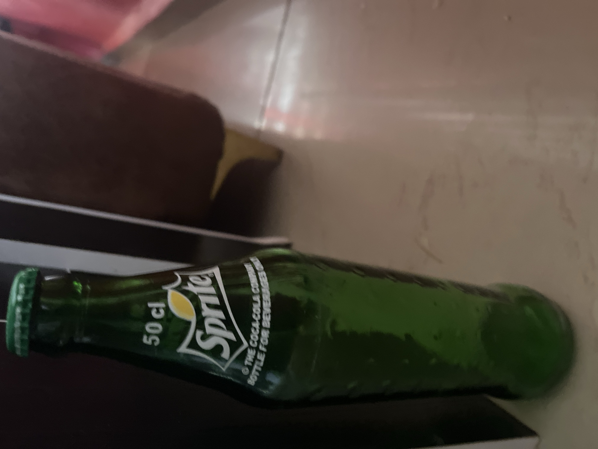 A green glass bottle of Sprite is lying on its side on a dark surface. The bottle has a green cap and the Sprite logo is visible on the label, along with the text "50 cl" and "© THE COCA-COLA COMPANY BOTTLE FOR BEVERAGES ONLY". The background is blurred, but appears to be an indoor setting with warm lighting. There are hints of pink and brown in the background, possibly indicating furniture or fabric. The image is taken from a close-up perspective, focusing on the bottle. No people are visible in the image. The scene is likely in Jos, Nigeria, given the provided location context.