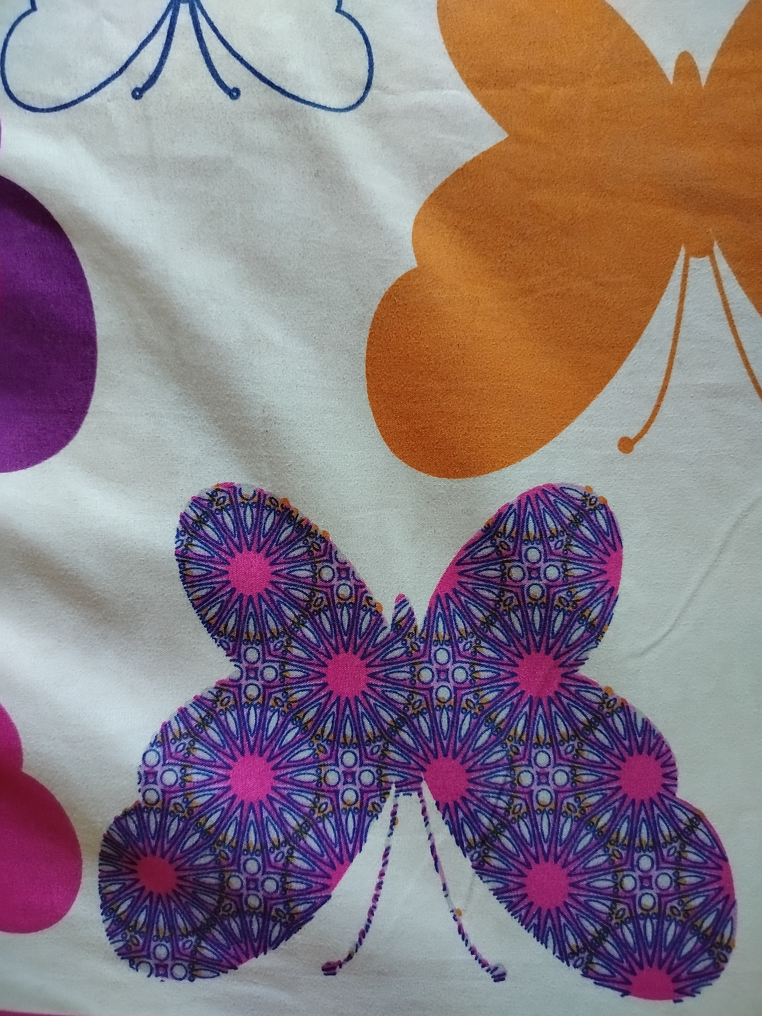 The image shows a close-up view of fabric with a butterfly pattern. The fabric is white and features several butterflies in various colors and styles. In the upper left portion of the image, there is a blue outline of a butterfly. To the right of that, a large, solid orange butterfly dominates the upper right quadrant. In the center of the image, a detailed butterfly is depicted with intricate patterns in shades of purple, pink, and blue, resembling stained glass or mosaic work. The fabric appears to be creased and textured, indicating it is likely a textile such as a bedsheet or curtain. The specific location in Bendet, Indonesia is not discernible from the image itself.