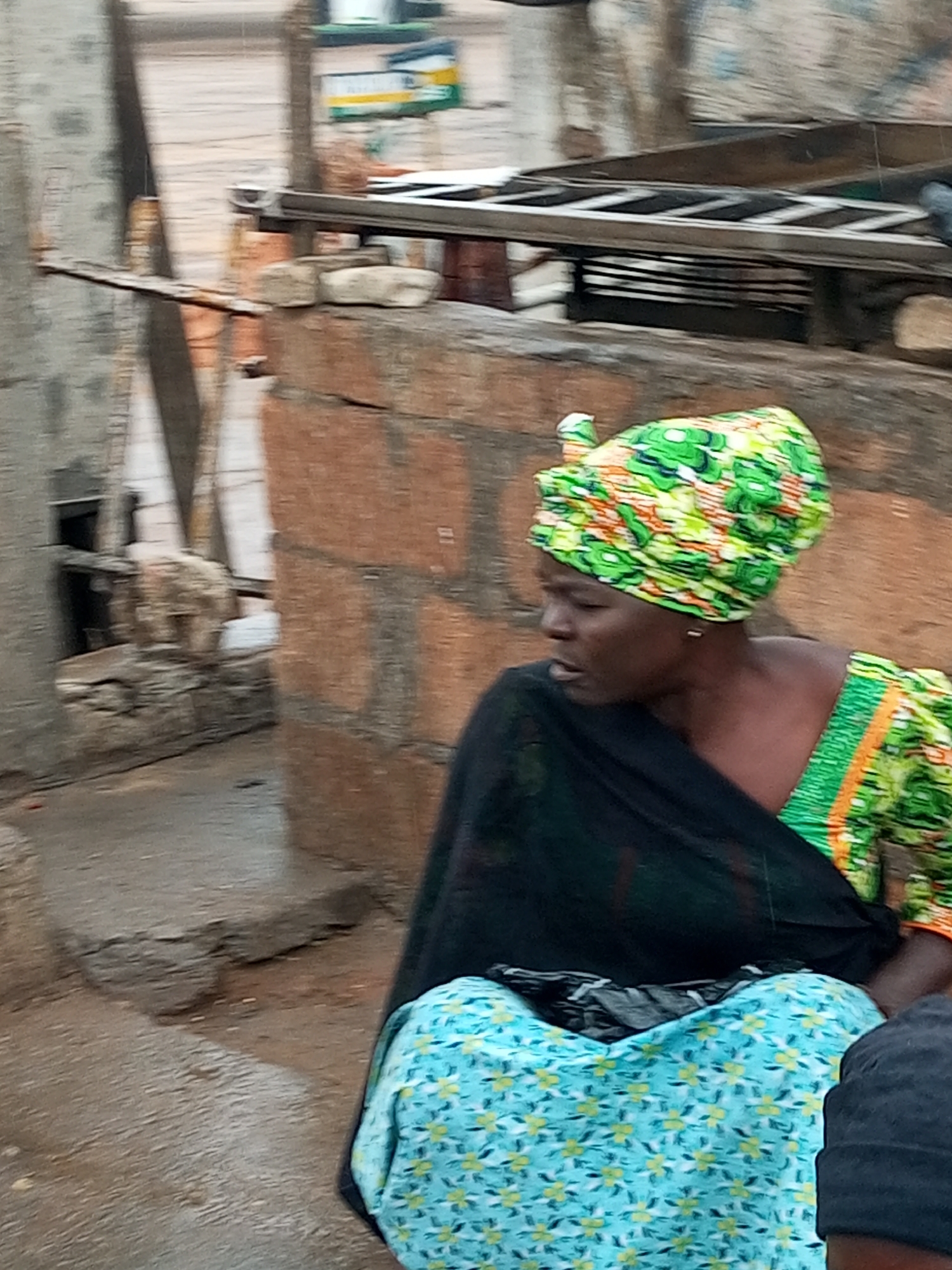 The image depicts a woman sitting in an outdoor setting, likely in Tempure, Nigeria. She is wearing a vibrantly patterned headscarf in green, yellow, and orange, and a matching patterned top with green and orange trim. A dark, possibly black, shawl is draped over her shoulders and torso, partially covering a light blue patterned fabric that appears to be a skirt or dress. Her expression is neutral, and she is looking downwards and to the left.
The background is composed of rough, reddish-brown brickwork, suggesting a rustic or unfinished structure. To the left of the woman, a portion of a grey concrete structure is visible, possibly part of a building or wall. In the upper part of the image, a metal ladder-like structure is precariously placed across the brickwork. The ground appears to be a mix of dirt and possibly paved surfaces, with some debris and uneven textures. The overall environment suggests a somewhat utilitarian or undeveloped urban area. There is no clear indication of specific actions or interactions, other than the woman's seated posture.