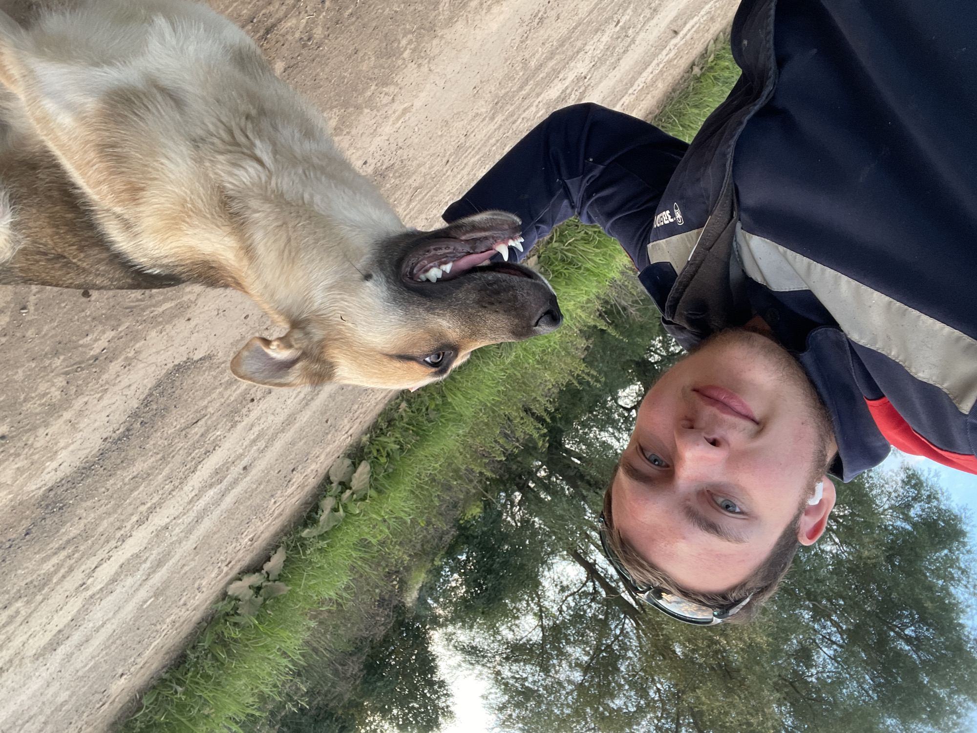 A young man with blue eyes and short brown hair, wearing a dark blue jacket with gray reflective stripes and white earbuds, is taking a selfie with a light brown and tan dog. The dog is looking towards the camera with its mouth open, showing its teeth and pink tongue. They are outdoors in a rural area. The ground is a dirt path, and to the left is a grassy embankment with trees. The sky is visible in the background. The image was taken in Panteleevka, Russia.