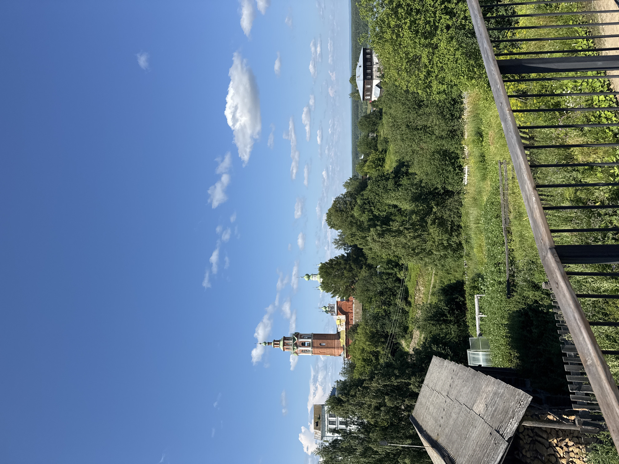 This is a photograph of the city of Cherdyn, Russia, taken from a high vantage point. In the foreground, a wooden railing with black metal supports overlooks a lush green landscape. To the left, the roof of a rustic building is visible, with a stack of firewood piled nearby. Beyond this, a greenhouse with a white frame is situated in a field of tall grass and wildflowers.
Dominating the mid-ground are several buildings, including a prominent red brick bell tower with green accents and a steeple. Adjacent to it are more structures, some with green domes, typical of Russian Orthodox churches. A white building with multiple windows stands to the left of the bell tower. The landscape is densely wooded with various shades of green trees.
In the background, a wider view of the city and surrounding countryside unfolds. A large, dark-roofed building with multiple windows is visible on a hill to the right. The horizon is dotted with more trees and distant buildings, under a bright blue sky with scattered white clouds. The overall impression is one of a serene, sunlit day in a historic Russian town.