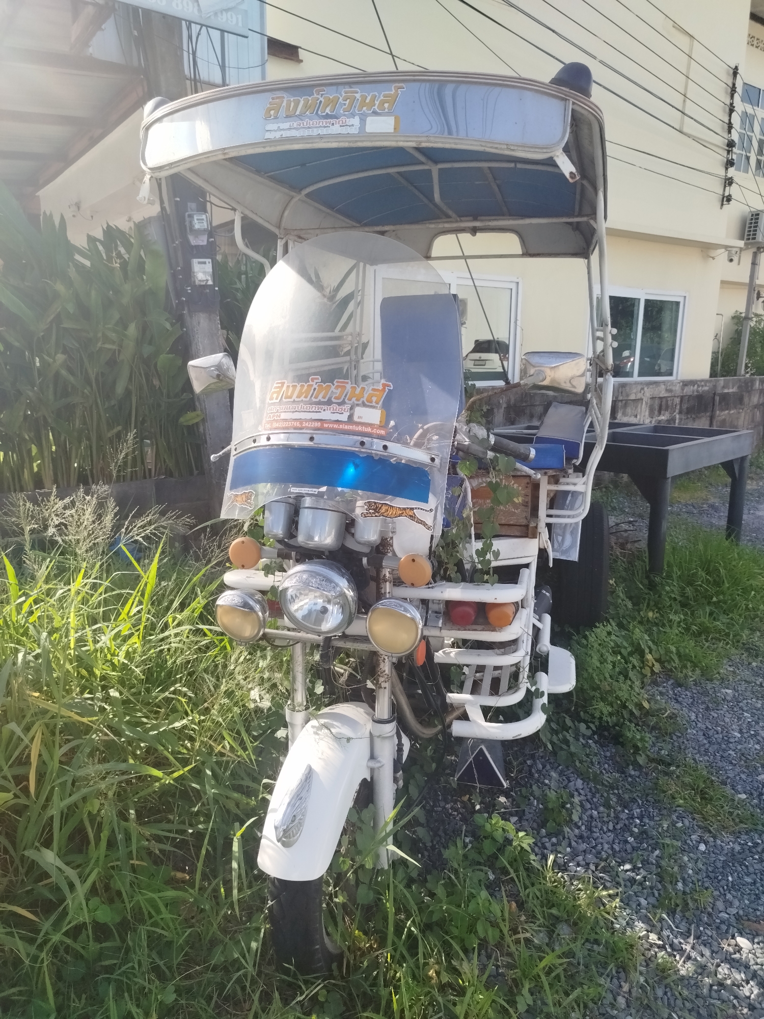 A white, three-wheeled vehicle, commonly known as a tuk-tuk, is parked on a gravel surface amidst tall green grass and foliage. The tuk-tuk has a white body with blue accents on its canopy and windshield. It features multiple lights on the front, including two large circular headlights and two smaller oval-shaped lights. The vehicle is equipped with a transparent windshield that displays some Thai writing and logos. A single seat, also with blue accents, is visible behind the driver's area. Behind the tuk-tuk, a dark grey, rectangular structure resembling a table or bench on legs is partially obscured by vegetation. In the background, a light yellow building with windows and electrical wires is visible. The scene appears to be outdoors, in what could be a residential or commercial area within Baan Lamai, Thailand. There are no people or animals present in the image.