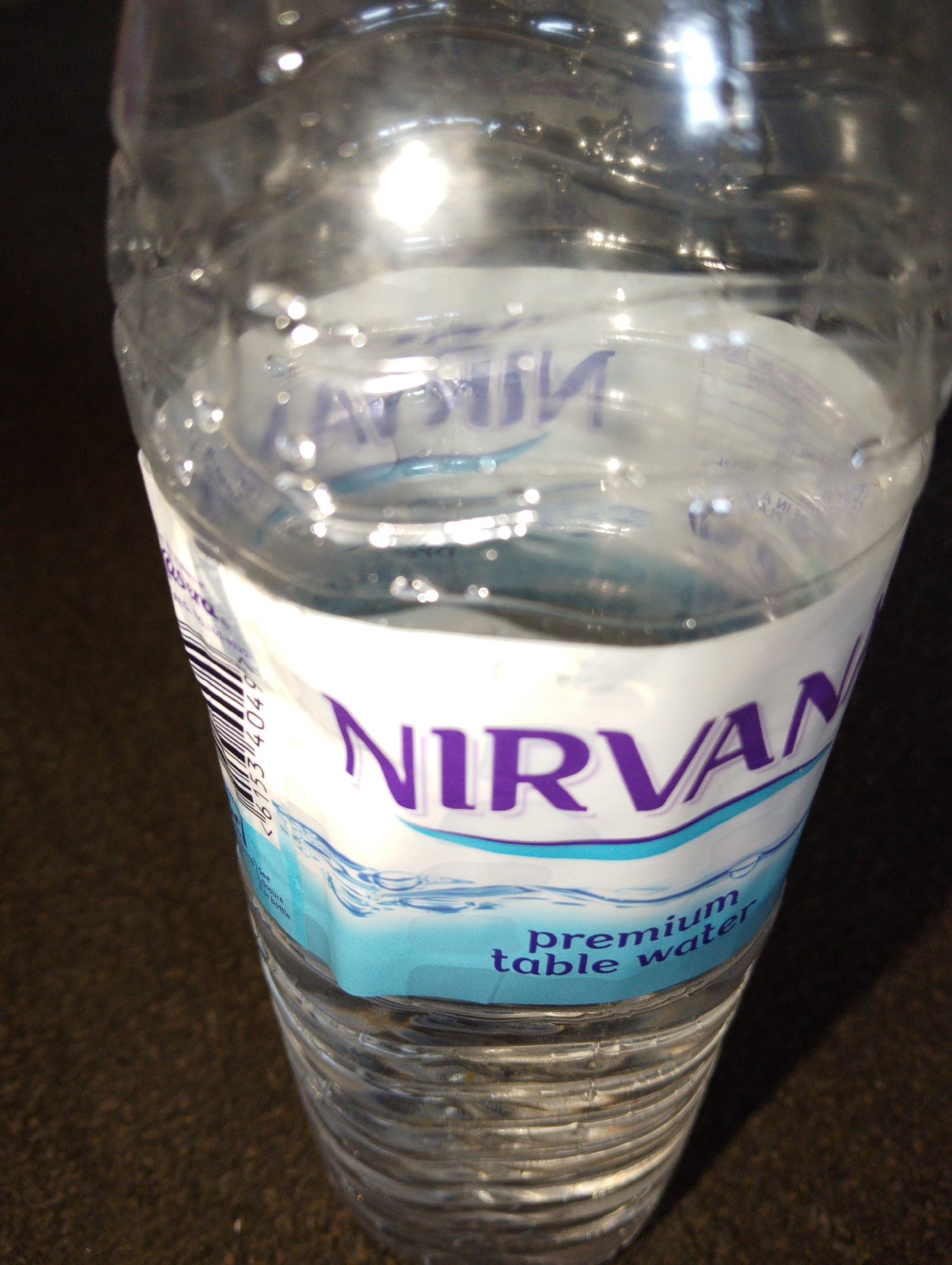 The image is a close-up shot of a clear plastic bottle of Nirvana premium table water. The bottle is partially filled with water, and condensation is visible on the exterior. The label on the bottle is white with purple lettering that reads "NIRVANA" and a blue band below with "premium table water" written in purple text. A portion of a barcode is visible on the left side of the bottle. The bottle is placed on a dark, textured surface that appears to be a table or countertop. The lighting suggests an indoor environment, possibly with overhead lighting reflecting off the bottle. There are no people or specific landmarks visible that would indicate a location within Lagos, Nigeria.