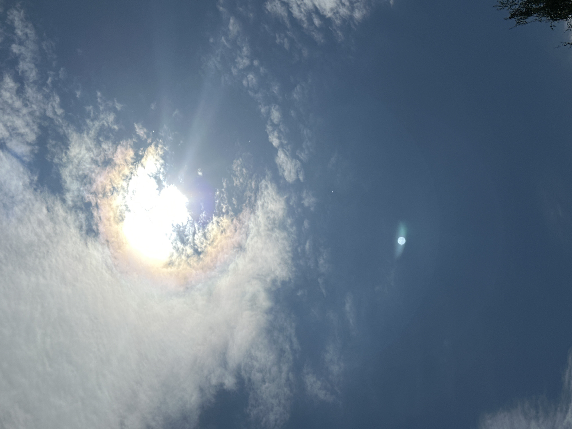 The image depicts a daytime sky with the sun partially obscured by thin clouds. A halo-like effect is visible around the sun. The sky is a gradient of blue, ranging from light to dark. It is impossible to determine the location as the city of Kokshetau, Kazakhstan. No people, actions or interactions are visible. The environment is simply the sky.