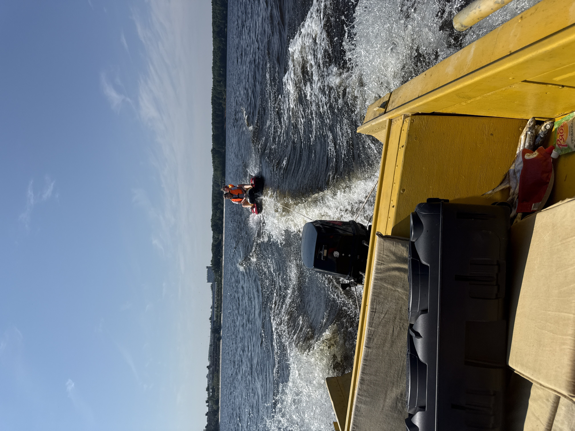 In Chaikovsky, Russia, on a sunny day, a yellow boat with a black outboard motor speeds across a dark, choppy river. The boat is leaving a foamy white wake. In the distance, two people are riding on an inflatable tube, also being pulled by the boat. They are wearing orange life vests. The riverbanks are lined with trees, and in the far distance, buildings are visible. The sky is a clear blue with a few wispy white clouds.