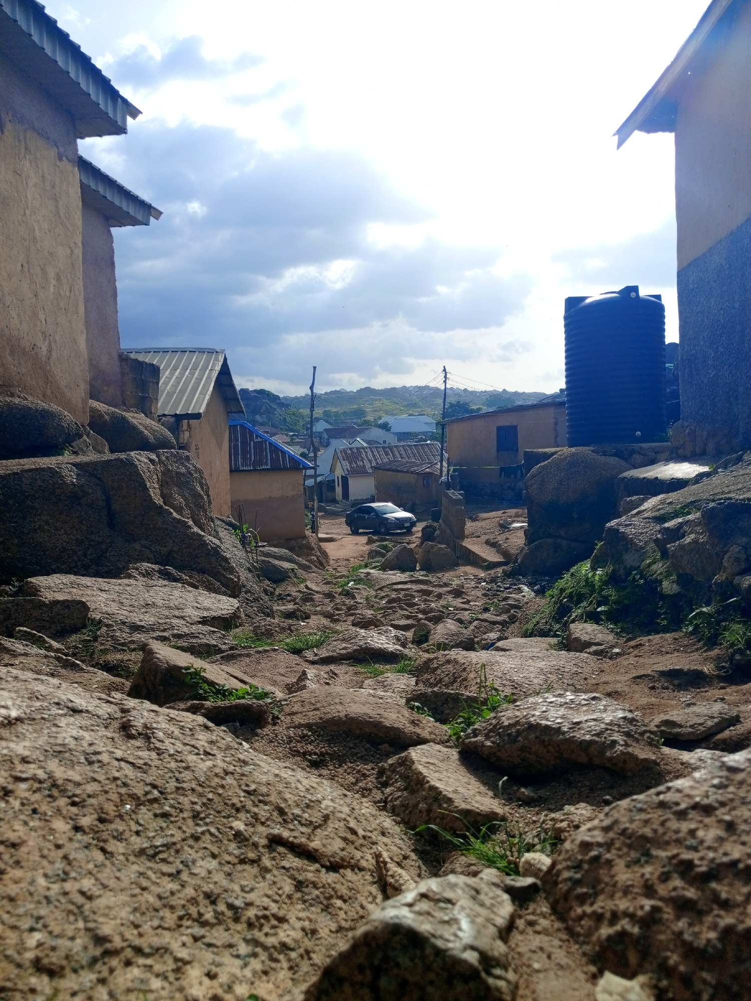 A low-angle shot depicts a rocky, uneven path leading through a village under a cloudy sky. The foreground is dominated by large, weathered rocks with patches of green grass growing between them. On either side of the path, simple buildings with corrugated metal roofs are visible. In the mid-ground, a dark-colored car is parked on the path, and a tall, dark blue water tank stands behind a building. In the background, more buildings and rolling hills can be seen under a bright, overcast sky. The overall scene suggests a rural or semi-rural environment with basic infrastructure.