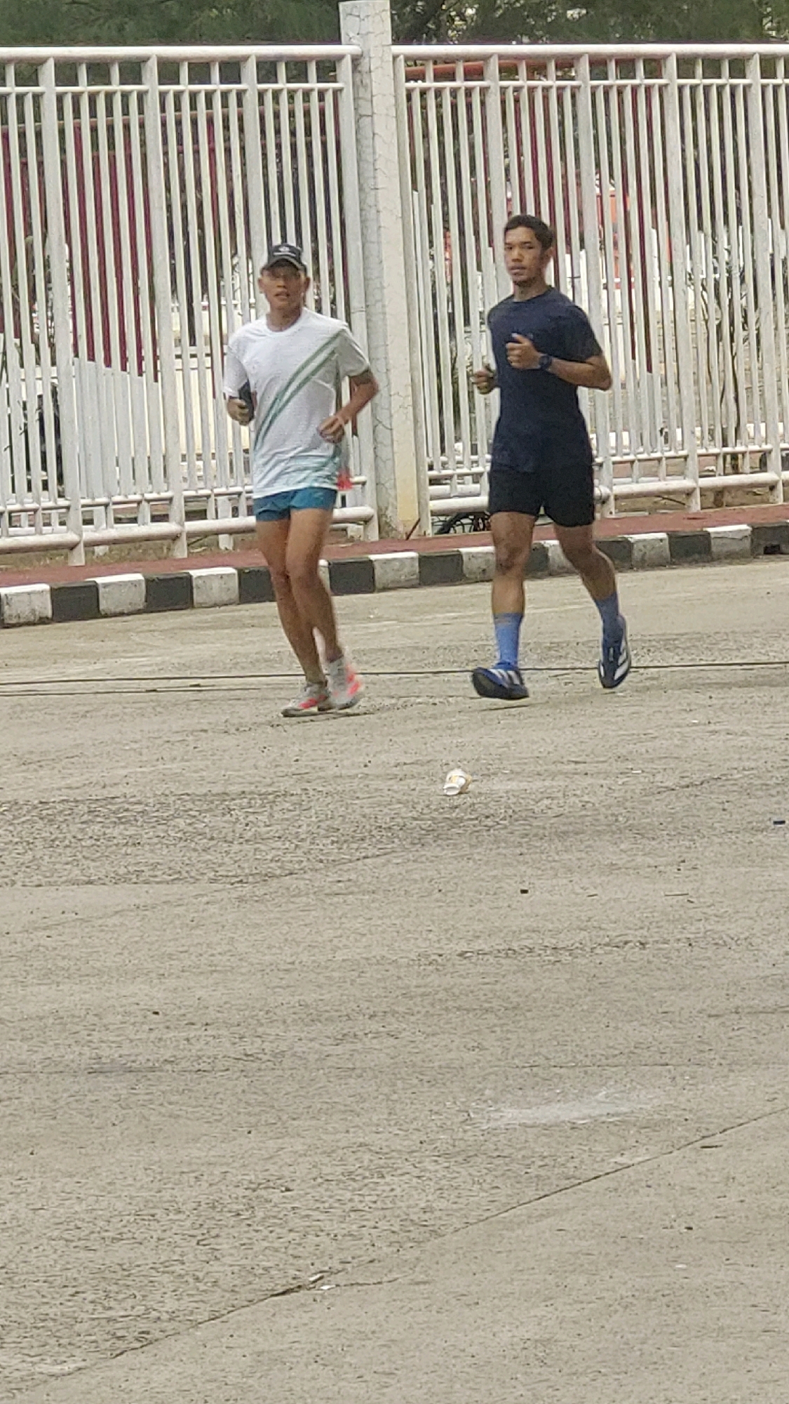Two men are jogging on a paved surface in what appears to be a park or outdoor recreational area in Cibinong, Indonesia, near a white metal fence. The man on the left is wearing a white t-shirt with green stripes, light blue shorts, and athletic shoes. He has a cap on and is holding something in his right hand. The man on the right is wearing a dark blue t-shirt, dark shorts, blue socks, and athletic shoes. He is looking forward and to the right. There is a white disposable cup on the ground between the two men. The environment is overcast with a slightly rough, grey paved surface. The fence behind the men is tall and white, with vertical bars and horizontal supports.