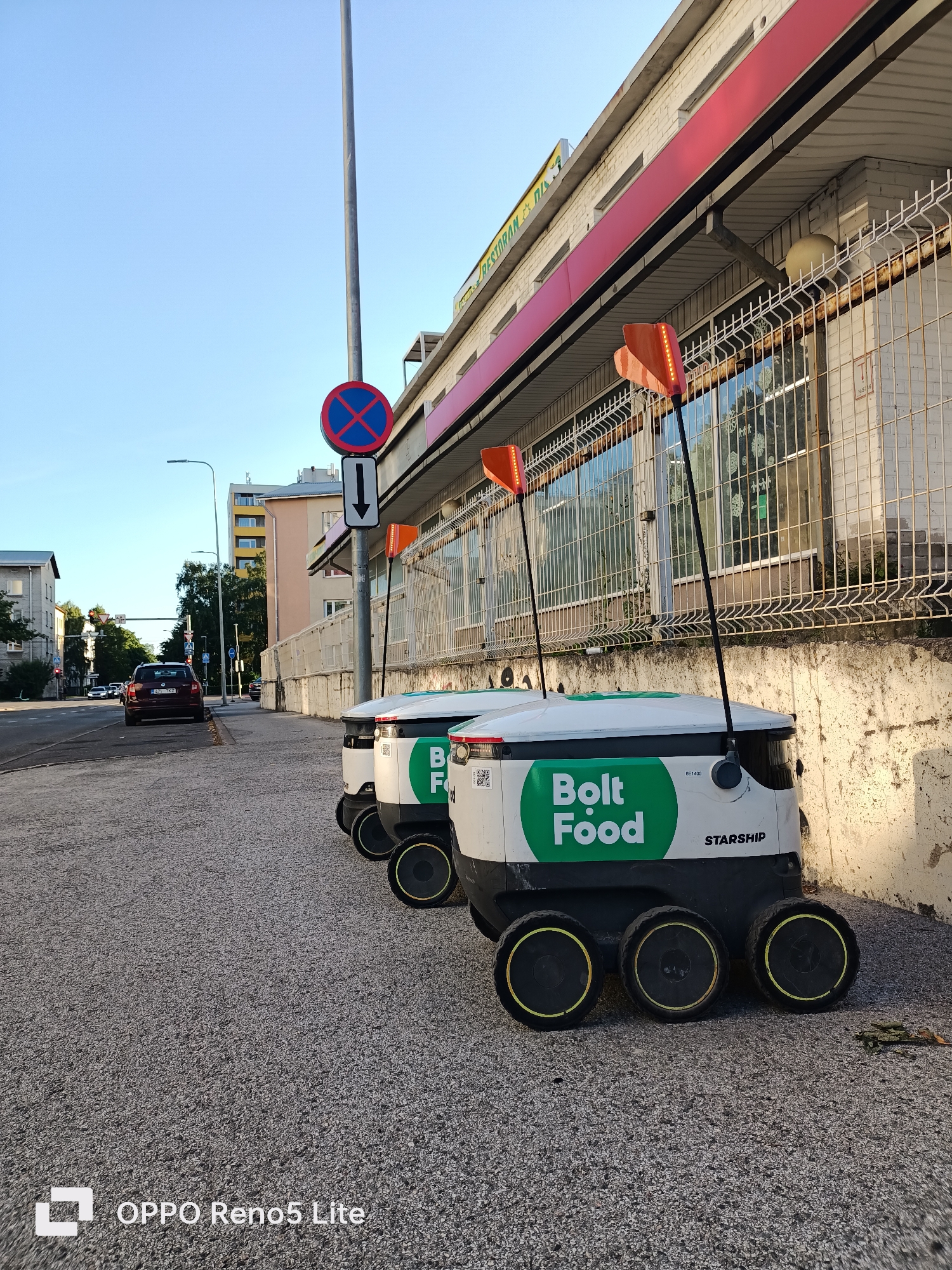 In Tallinn, Estonia, on a sunlit day, three autonomous delivery robots, branded with "Bolt Food" and "STARSHIP," are parked on a gravel sidewalk next to a building with a red awning. The robots are white with green circular logos and have orange flashing lights on poles extending from their tops. A "no parking" sign with an arrow pointing downwards is visible on a pole. Across the street, a dark-colored car is parked, and in the background, apartment buildings and trees line the street. The scene suggests a functional urban environment where delivery robots are being utilized.