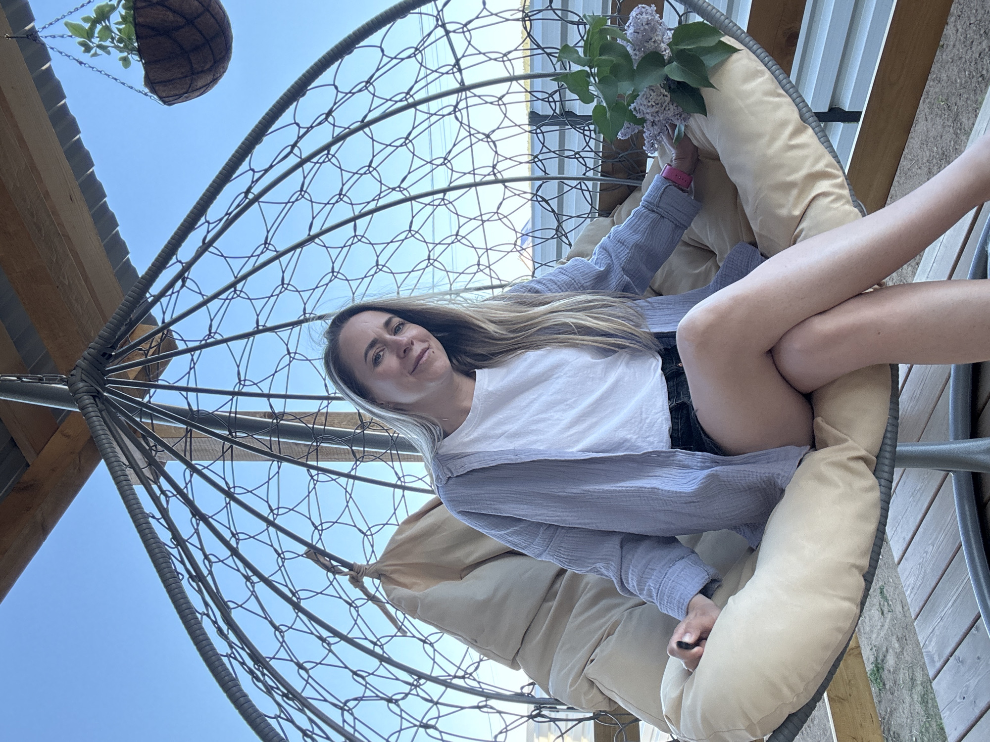 A woman is sitting in a hanging chair, possibly in a garden or outdoor setting. She is wearing a white t-shirt and a light blue button-down shirt. She has her legs crossed and is holding a bouquet of lilacs. The chair is made of a metal frame with a woven wire design and has beige cushions. A hanging basket with plants is visible in the upper left corner of the image. The background includes a blue sky, a wooden structure, and some greenery. There is no evidence to suggest that this image was taken in Chaikovsky, Russia.
