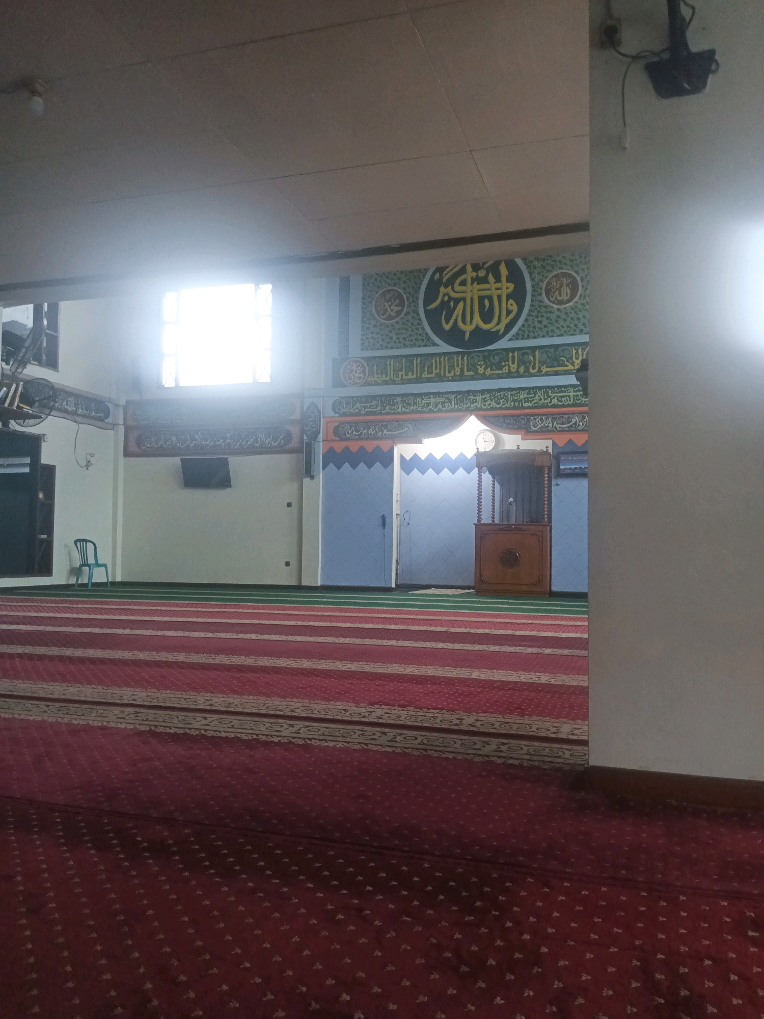 The image shows the interior of a mosque, likely in South Tangerang, Indonesia, based on the Arabic calligraphy prominently displayed on the wall. The foreground features a red carpet with a patterned design, indicative of prayer mats. Behind the carpeted area, a green carpeted floor is visible. Further into the mosque, a wooden podium stands against a wall decorated with intricate Arabic calligraphy and geometric patterns. A clock is mounted on the wall above the podium. To the left, a light blue chair is placed near a wall, and a television screen is mounted on the wall. The environment appears to be indoors, with artificial lighting and natural light streaming through a window. The scene is devoid of people, suggesting it's either before or after prayer times, or during a quiet period.